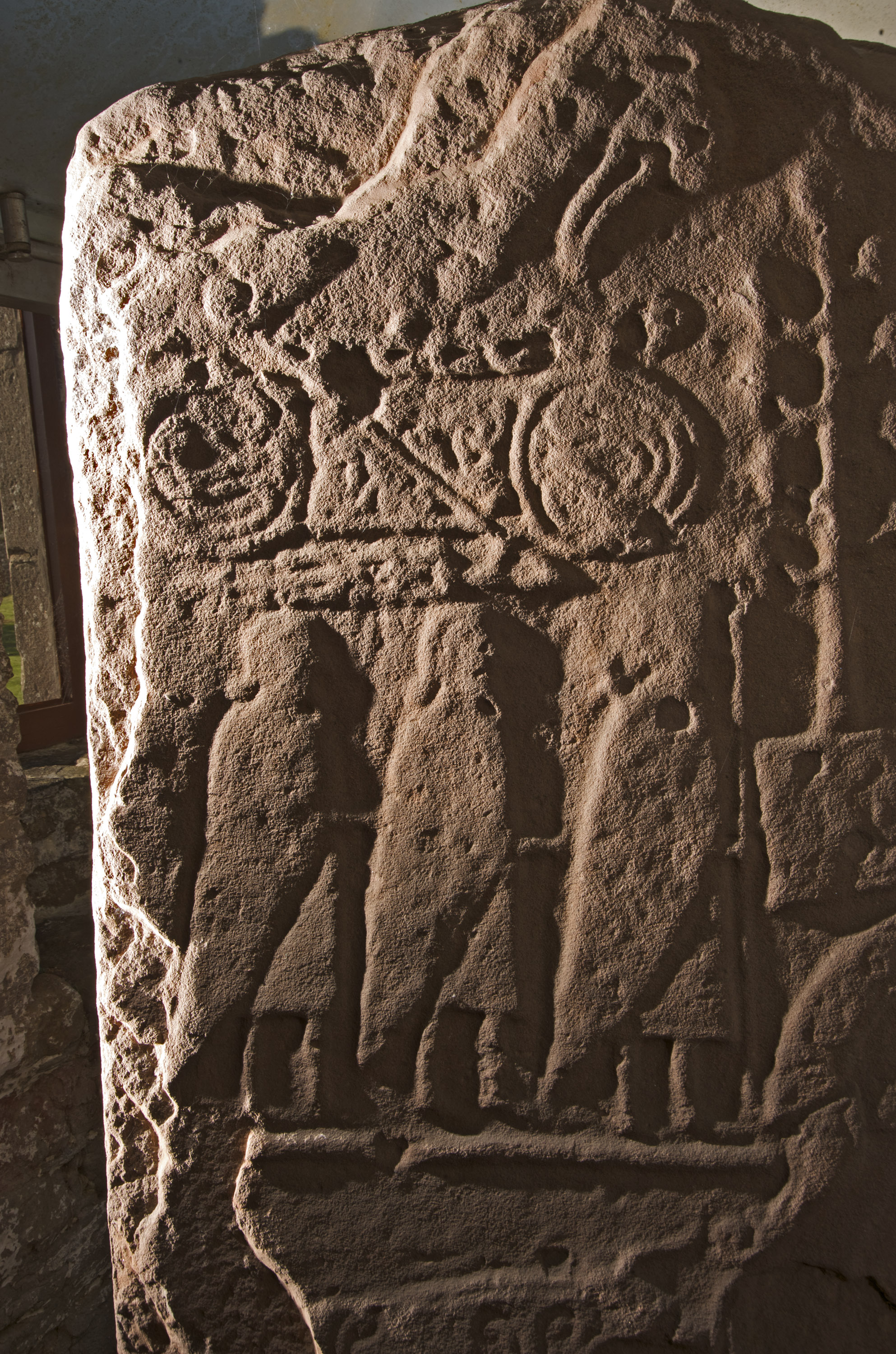 Close‑up view of the Eassie Pictish sculptured stone showing a worn but detailed carved panel. The upper section features a pair of large circular symbols connected by a horizontal bar with additional abstract shapes above. Below this are three robed standing figures carved in low relief. The sandstone surface is rough and weathered, with directional lighting emphasising the carved lines and textures. 