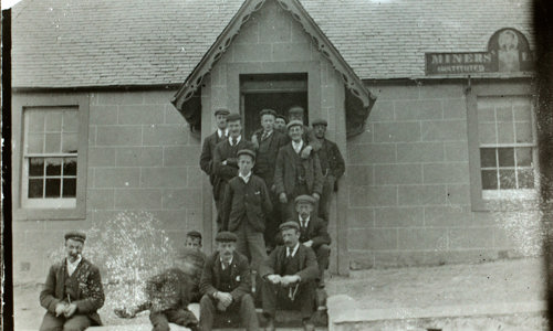 A black and white photo shows 14 men dressed in suits with black boots and flat caps. They sit and stand on the steps of a stone building with a slate roof.