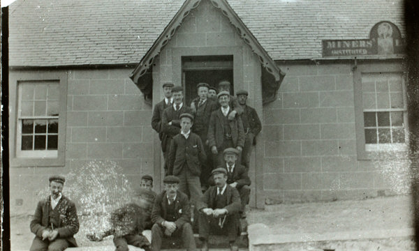 A black and white photo shows 14 men dressed in suits with black boots and flat caps. They sit and stand on the steps of a stone building with a slate roof.