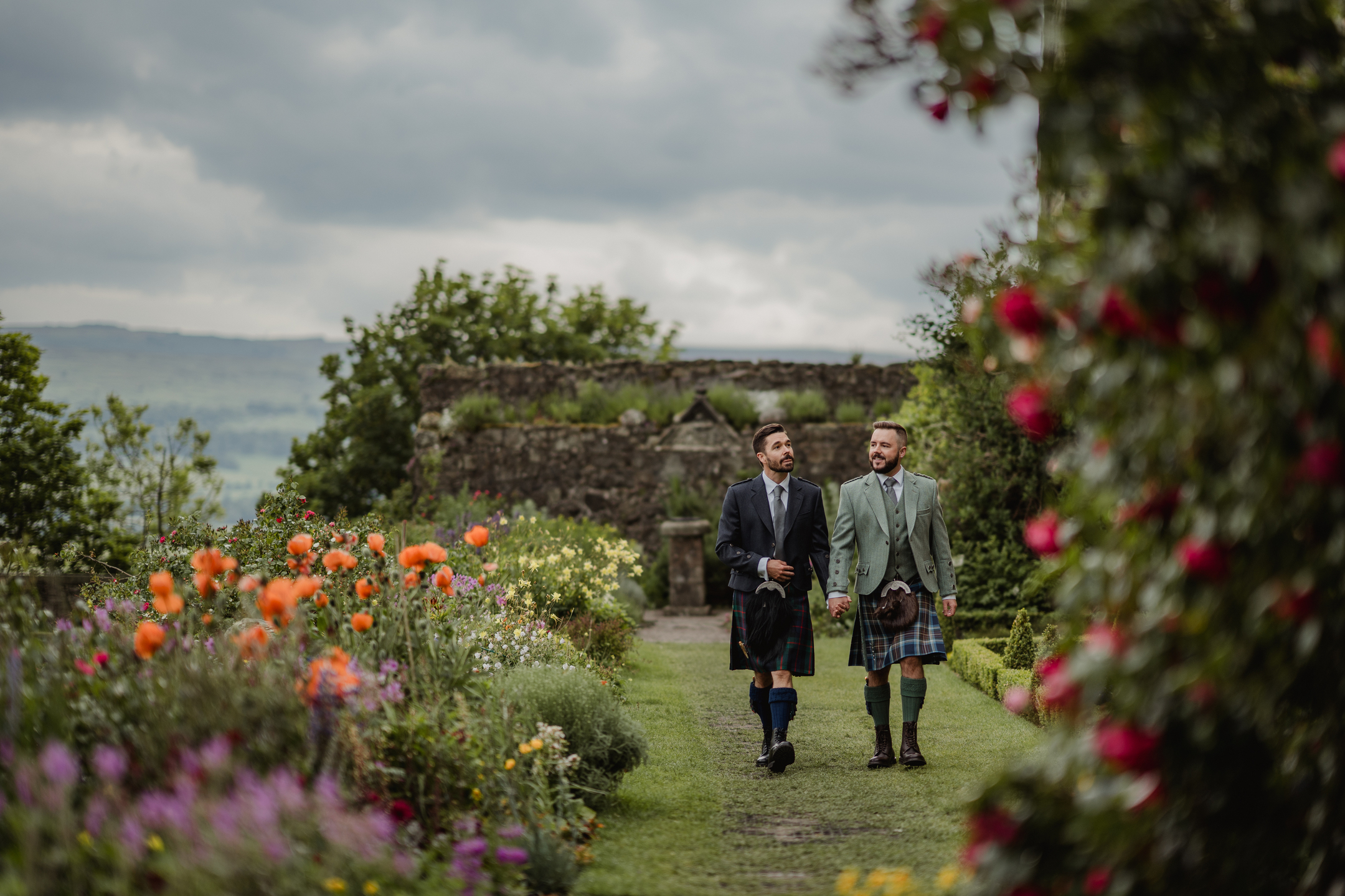 Two Grooms walking hand in hand in hand through a garden filled with colourful flowers