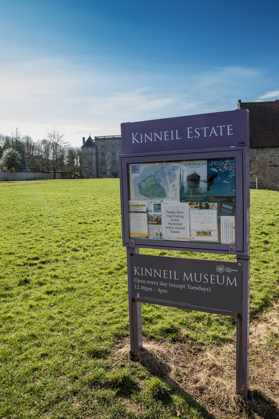 A noticeboard sits on a grass landscape, it reads 'Kinneal Estate', with notices pinned below it. Below is a sign to Kinneil Museum. There is a building in the background, and it is a sunny day. 