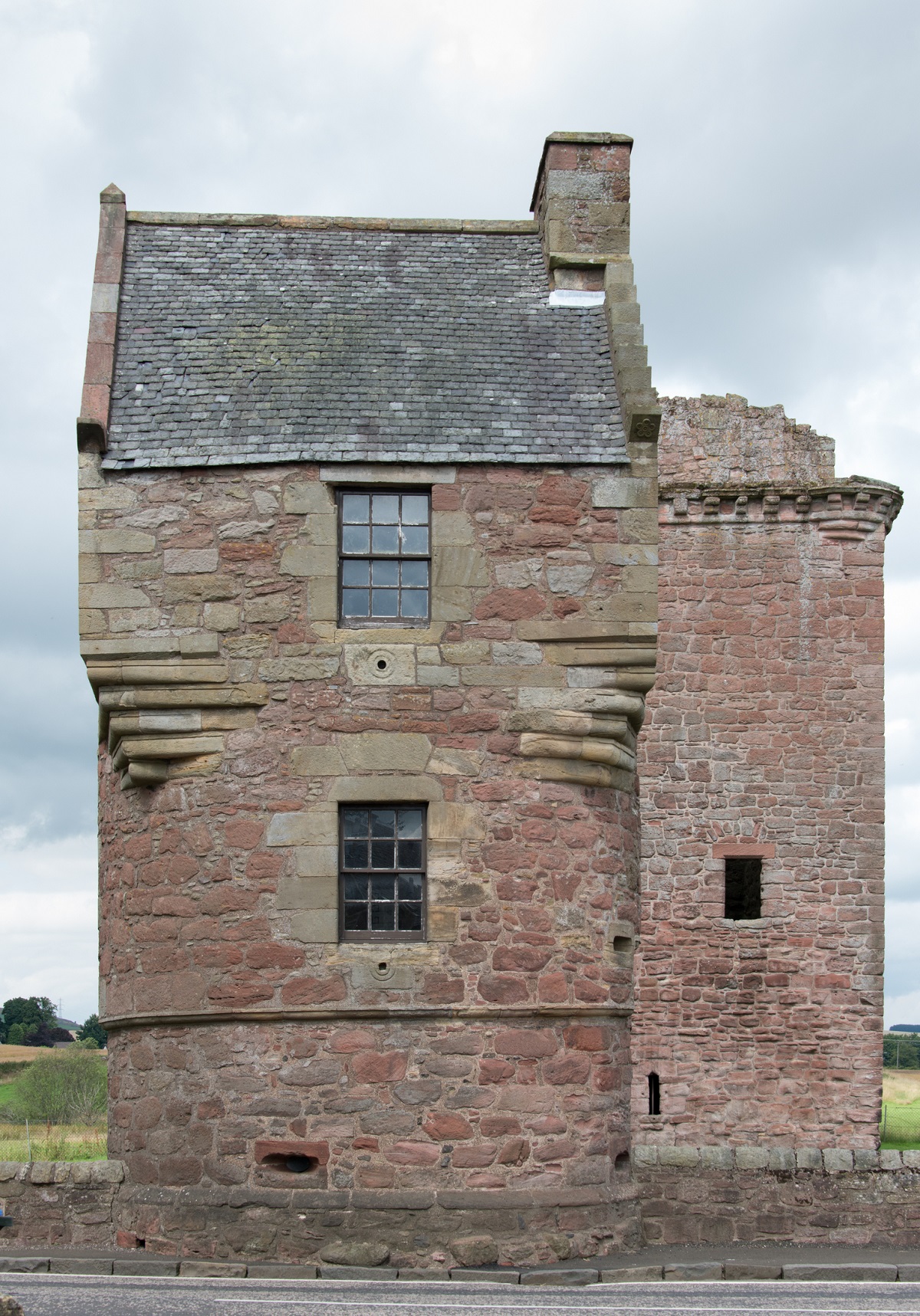 Front view of a rectangular stone tower at Burleigh Castle with small windows and a slate roof, with a second tower partially visible behind it under a cloudy sky.