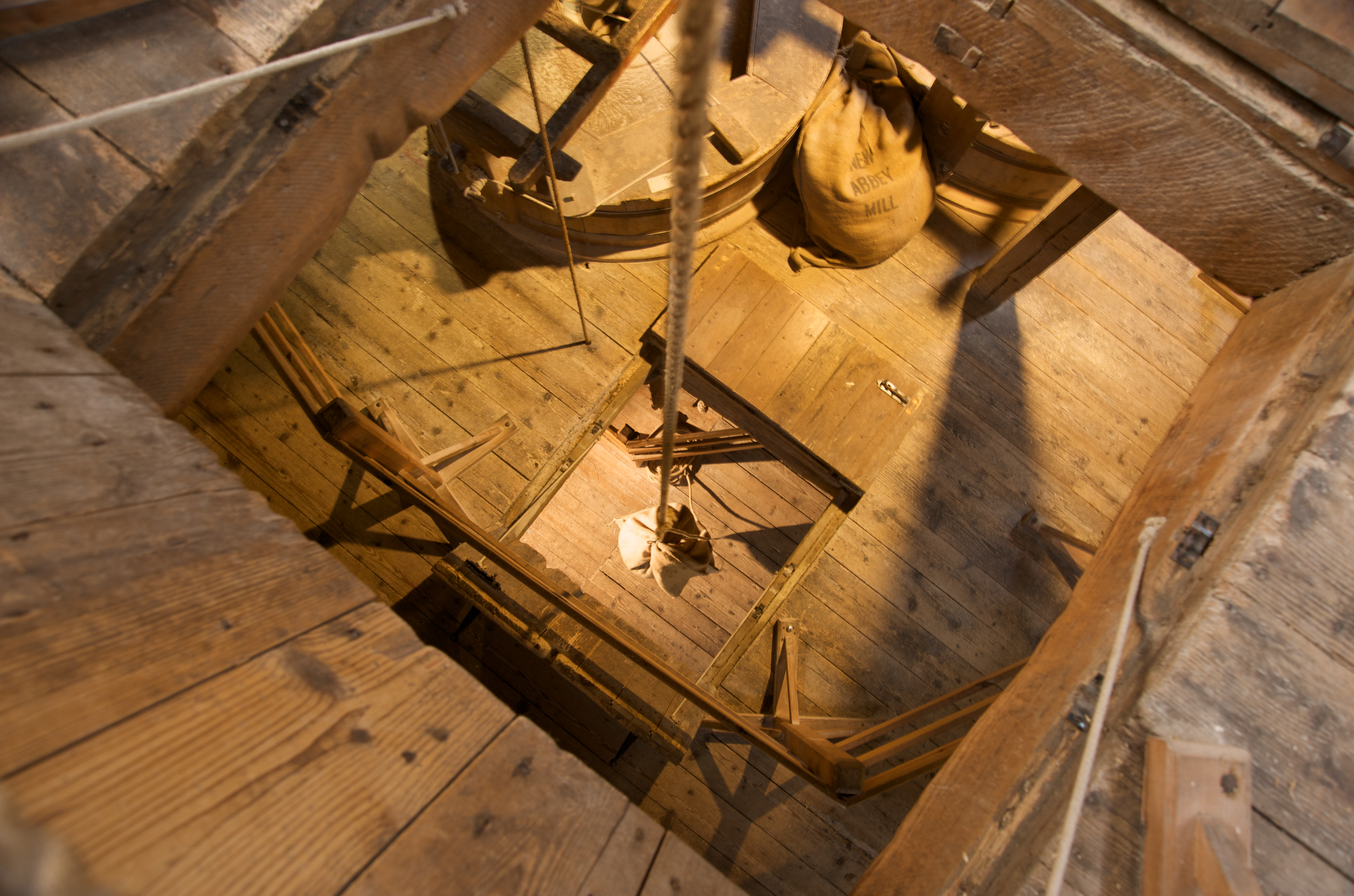 A view looking down through a square opening in the wooden mill floor, with a rope and pulley system hanging above and sacks and mill machinery surrounding the space.