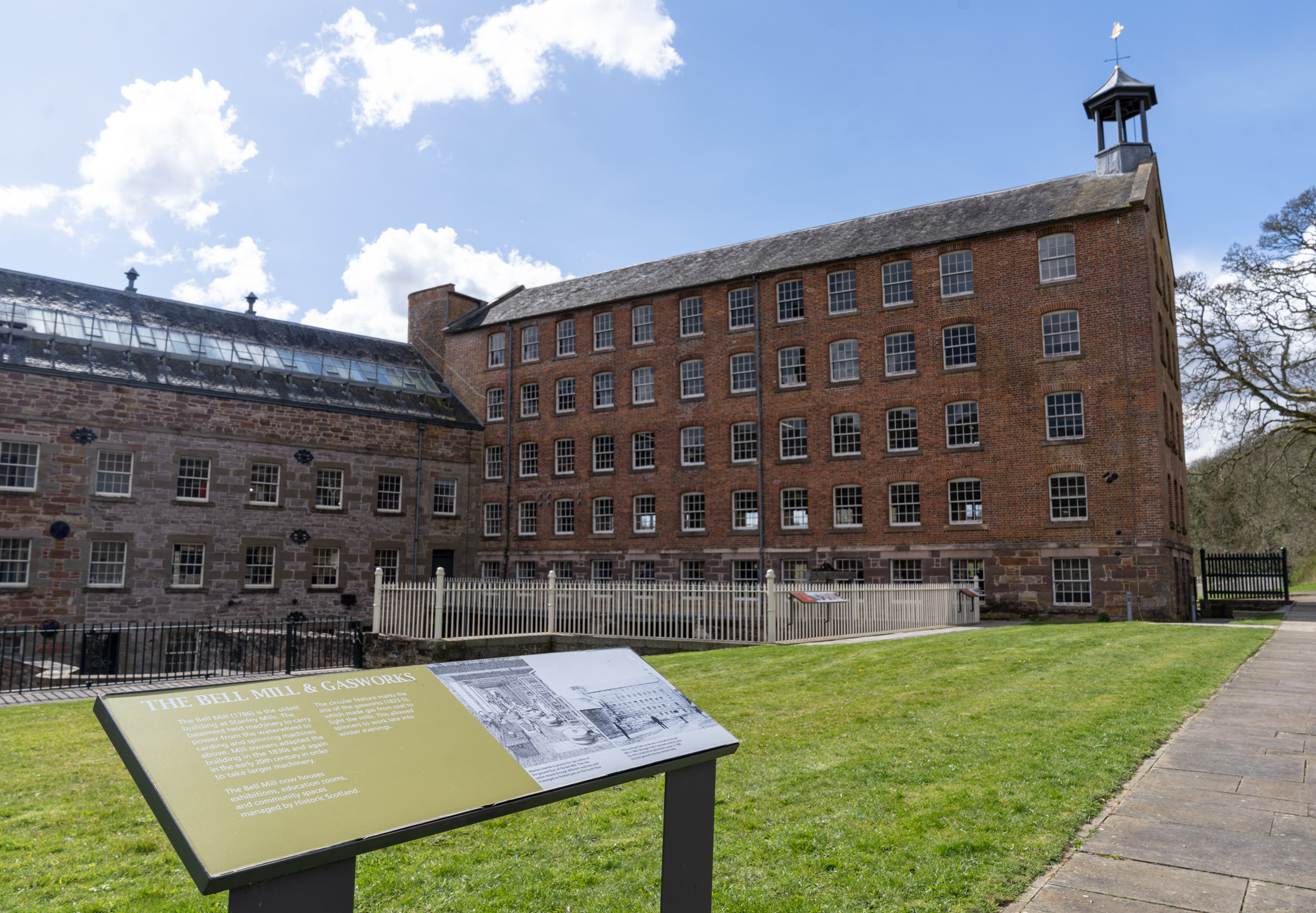 An outdoor interpretation panel about the Bell Mill and gasworks stands in front of tall brick mill buildings with rows of windows and a fenced grassy area.