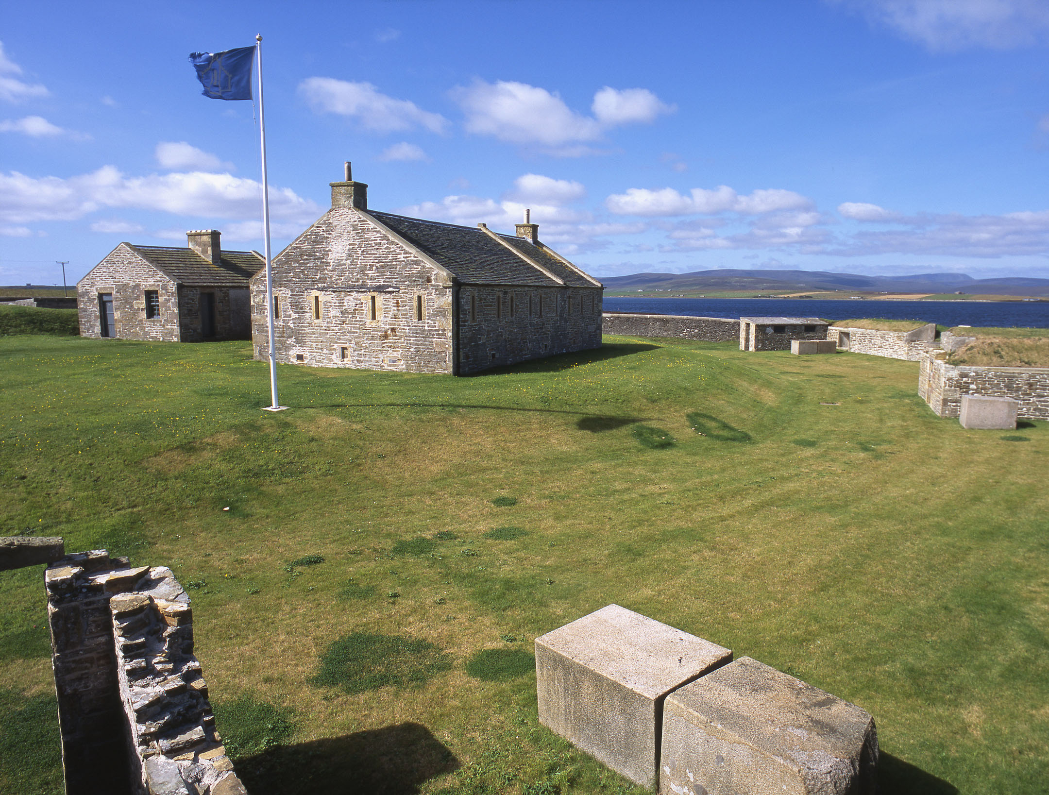 Stone barracks buildings with flagpole on grassy slope overlooking water.
