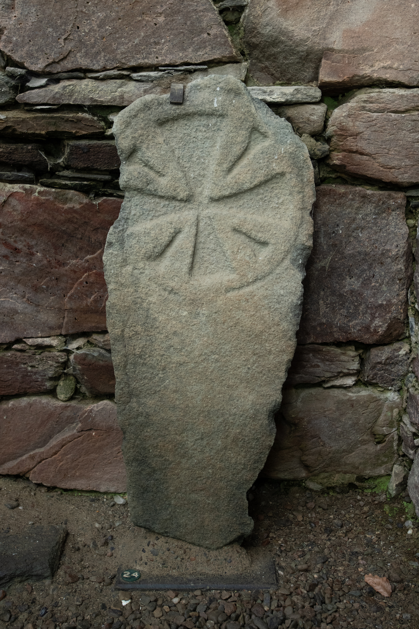 A rough, irregular grave slab carved with a wheel-like circular motif, mounted against a stone wall.