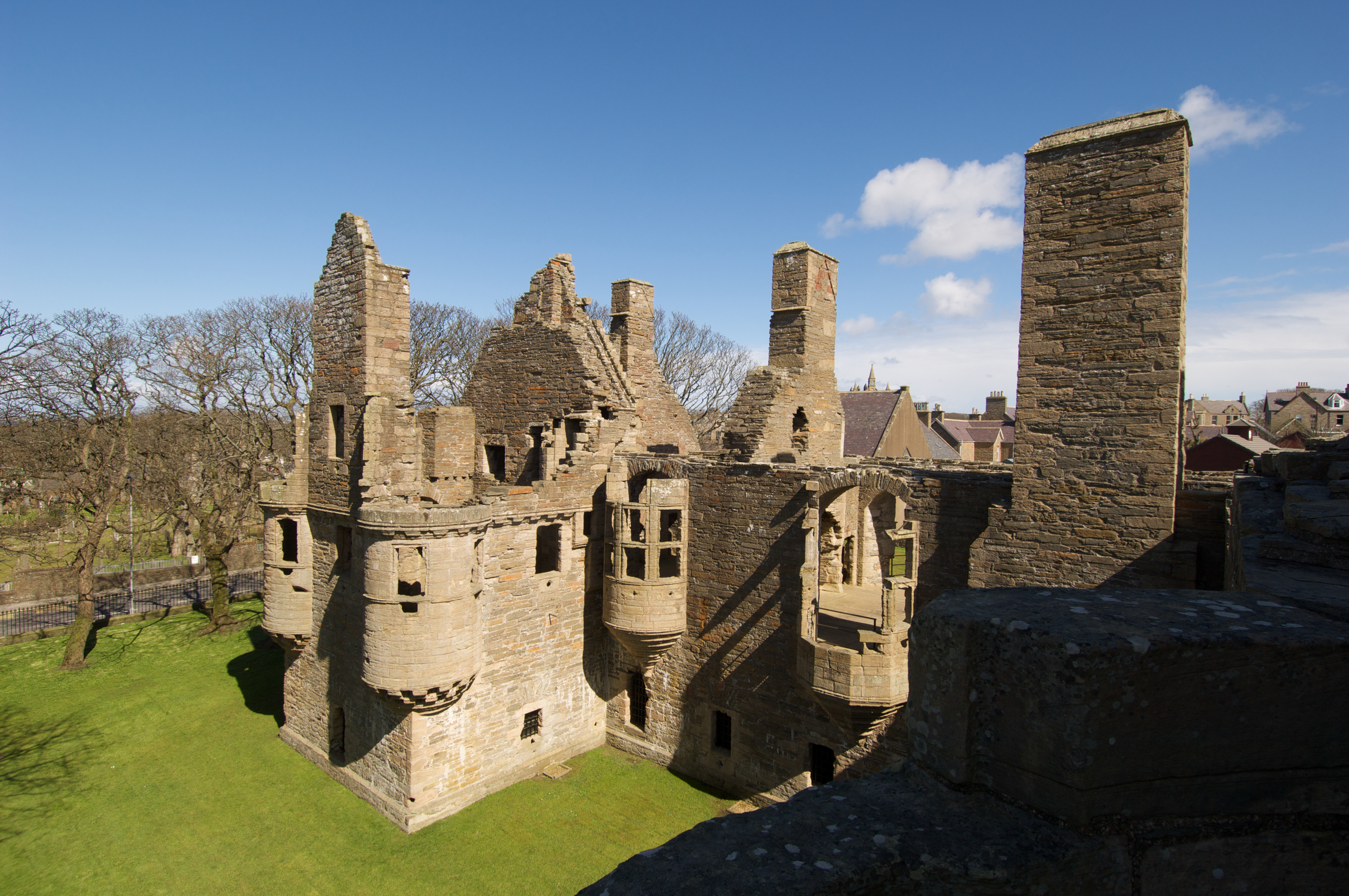An elevated view of a large stone palace ruin with tall chimneys, turrets, and partially collapsed walls, surrounded by grass and trees.