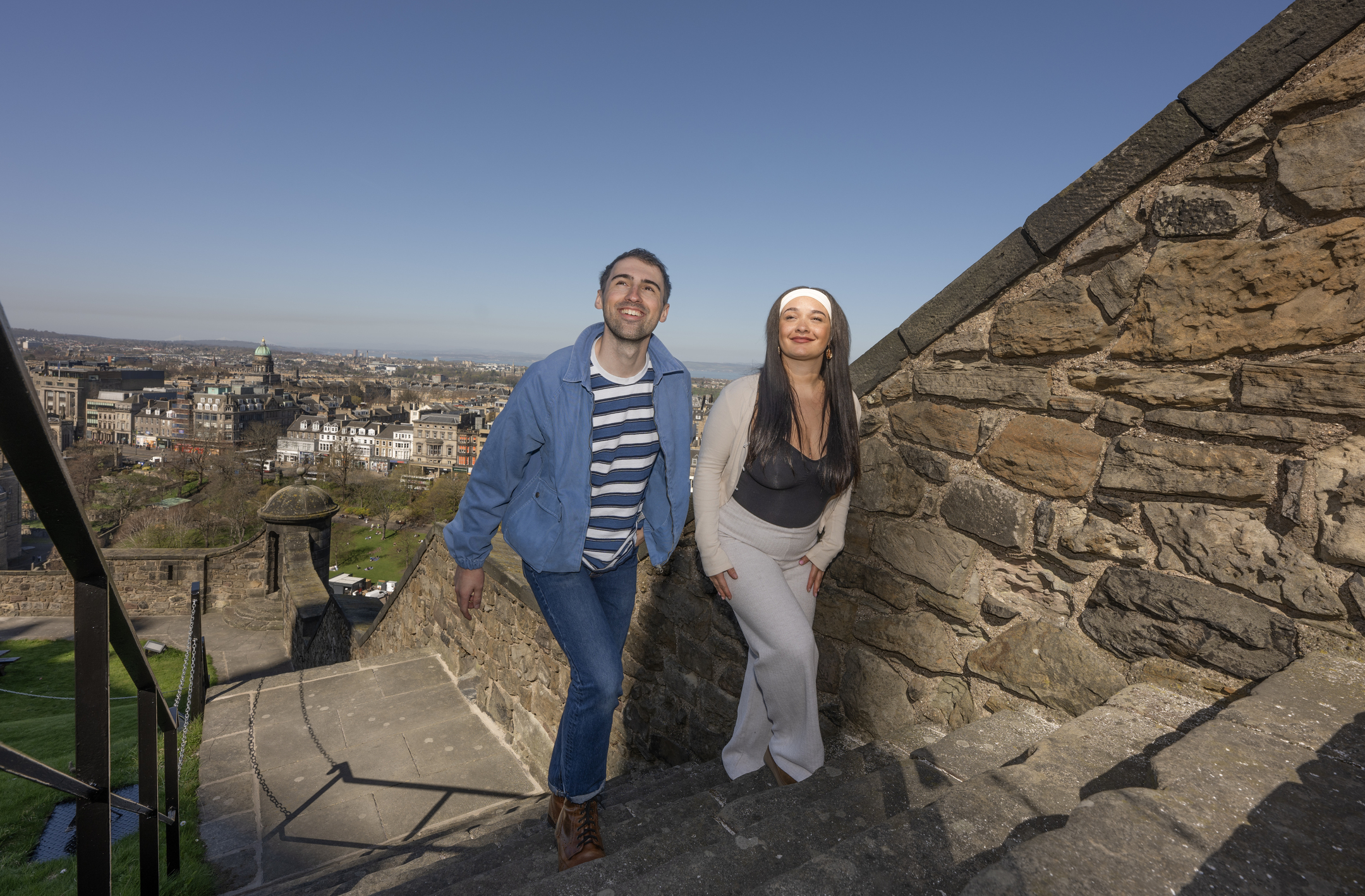 Two visitors climb a stone staircase at Edinburgh Castle on a bright, sunny day. There is a clear blue sky over the city behind them.