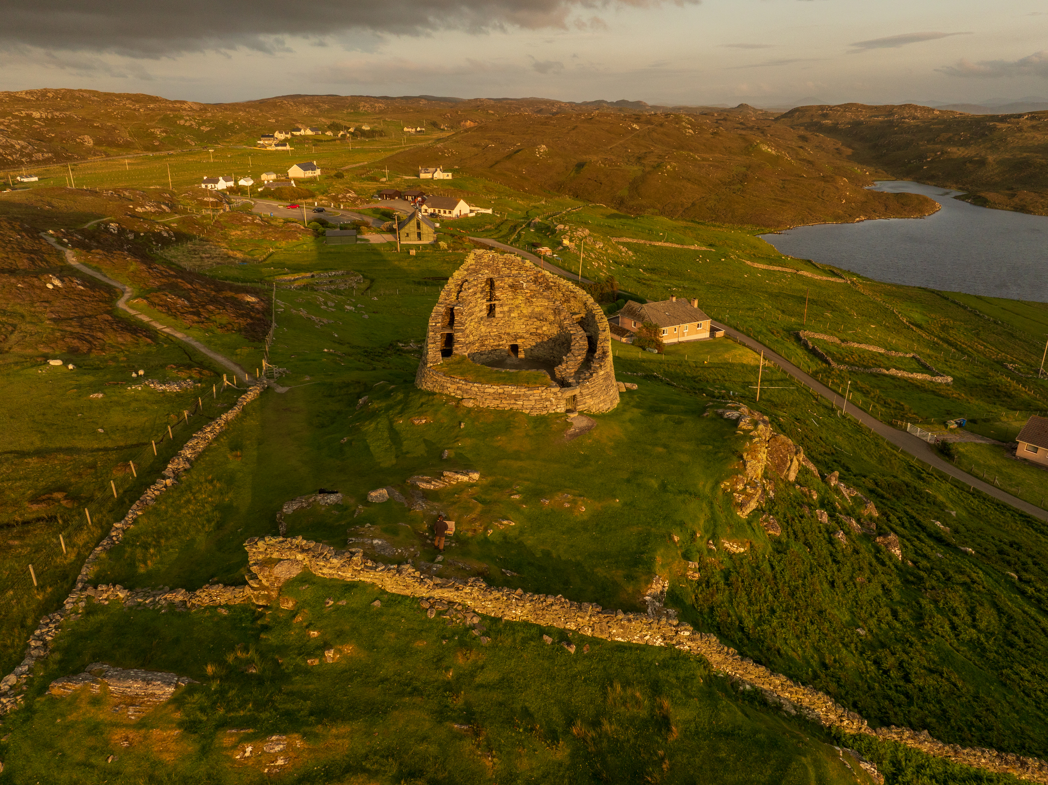 Aerial view of a stone ruin on a grassy hill overlooking a lake and rolling countryside at sunset.