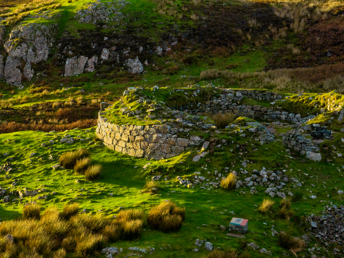 A circular stone wall, with a break where the stone has crumbled. There is grass surrounding it.