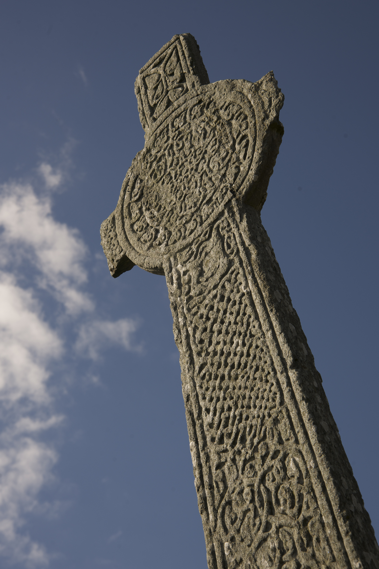 Low‑angle view of a tall carved stone cross showing detailed knotwork and circular patterns etched into its surface. The cross rises sharply against a deep blue sky with a few scattered clouds, emphasizing its height and textured stonework.