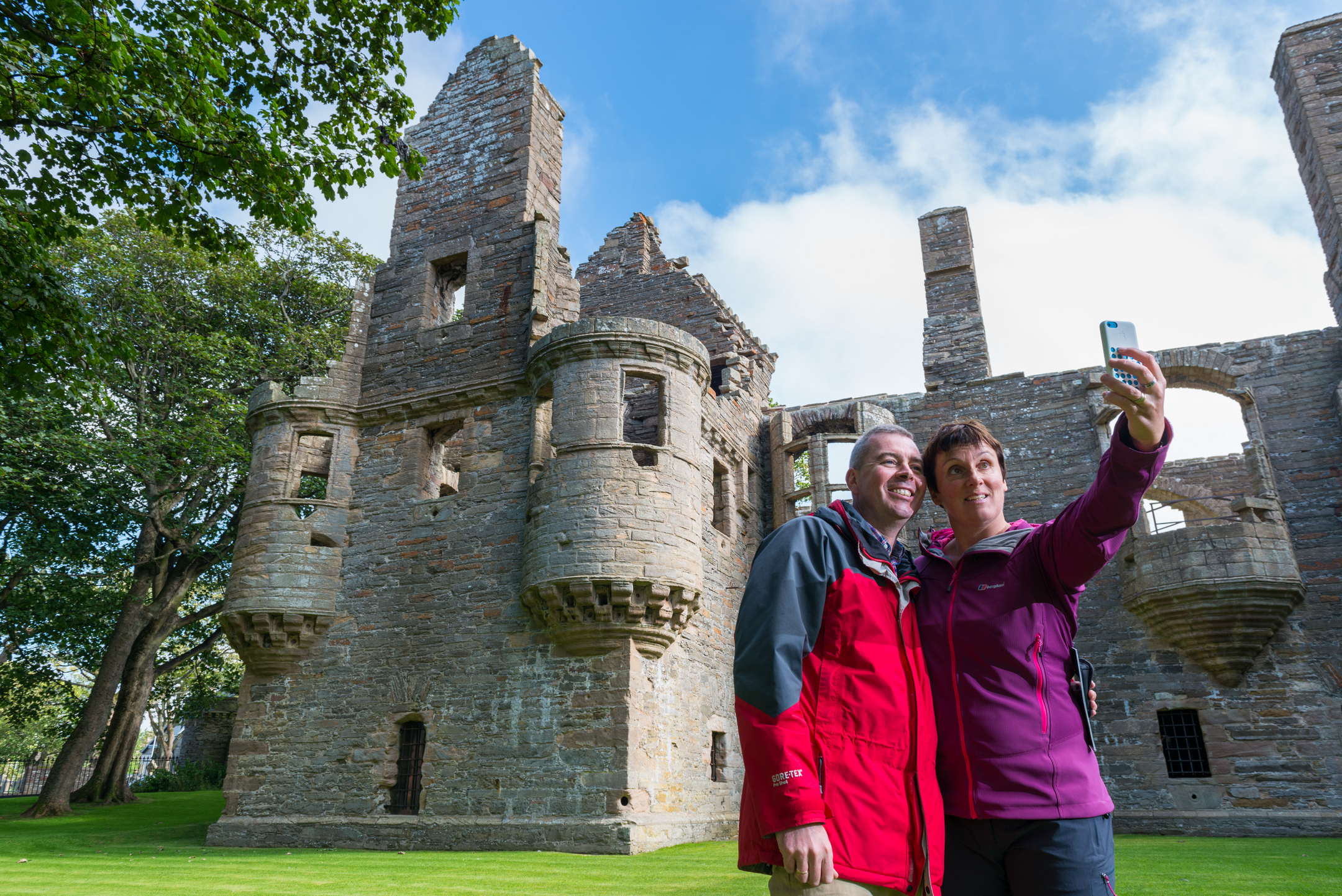Two people taking a selfie in front of a large stone palace ruin with towers and arched windows, set on a grassy lawn with trees nearby.