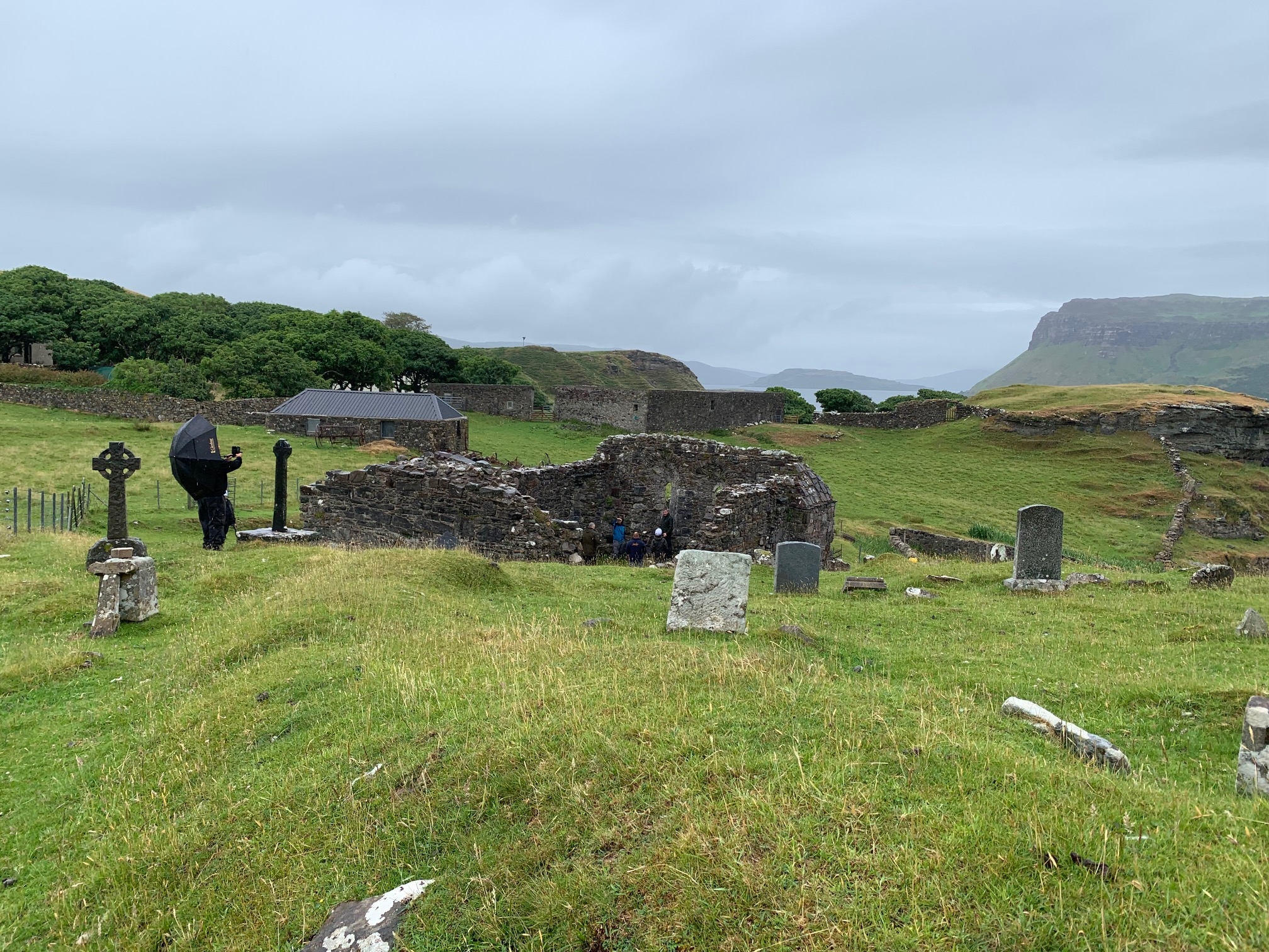 View of the chapel ruins from higher ground, looking across a graveyard with old headstones and a person holding an umbrella on a rainy day.