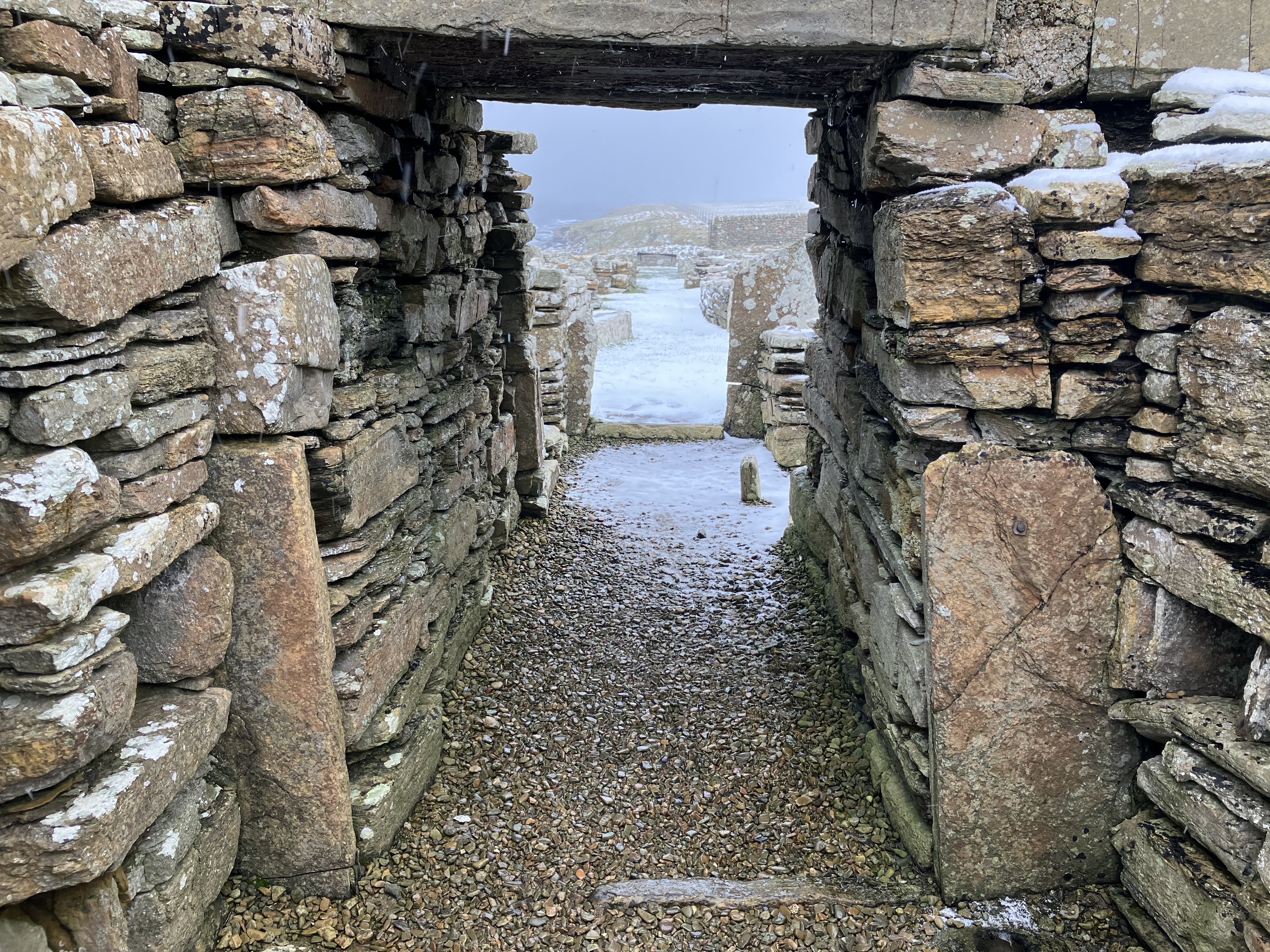 Interior view through a narrow stone passageway with stacked slabs forming walls and a small doorway leading to another chamber. Light filters through from outside.