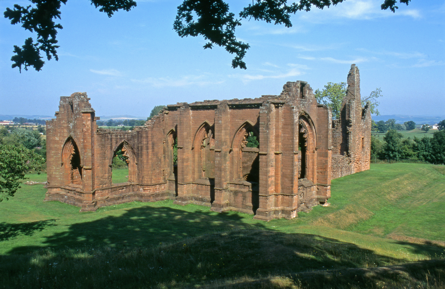 Side view of historic stone ruins, with multiple tall arches with detailing around the outside. Some of stone has crumbled, particularly at the top. It sits on a grassy landscape on a sunny day.