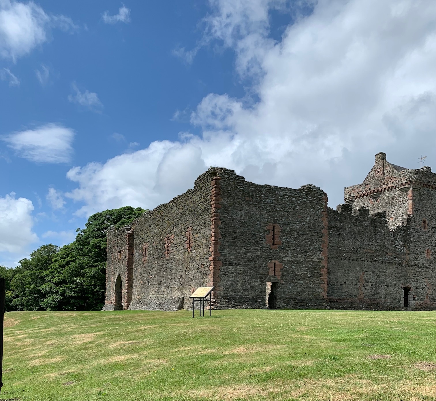 A large rectangular stone castle ruin with thick walls and narrow windows, set on a grassy lawn beneath a bright blue sky with scattered clouds.