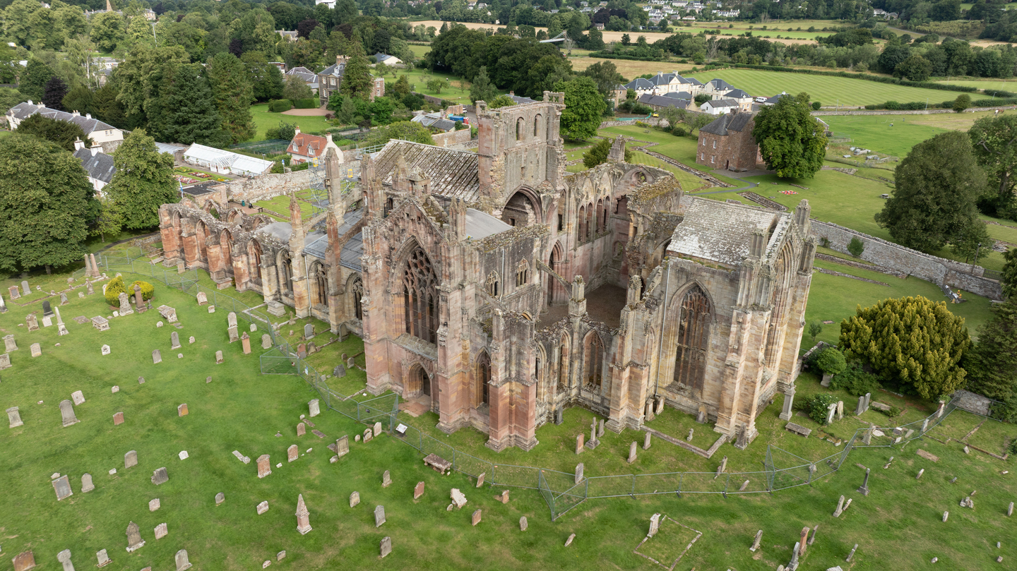 Aerial view of a historic, stone building, reminiscent of an abbey. Some of the stone is weathered. There is a graveyard surrounding it. and trees in the background.