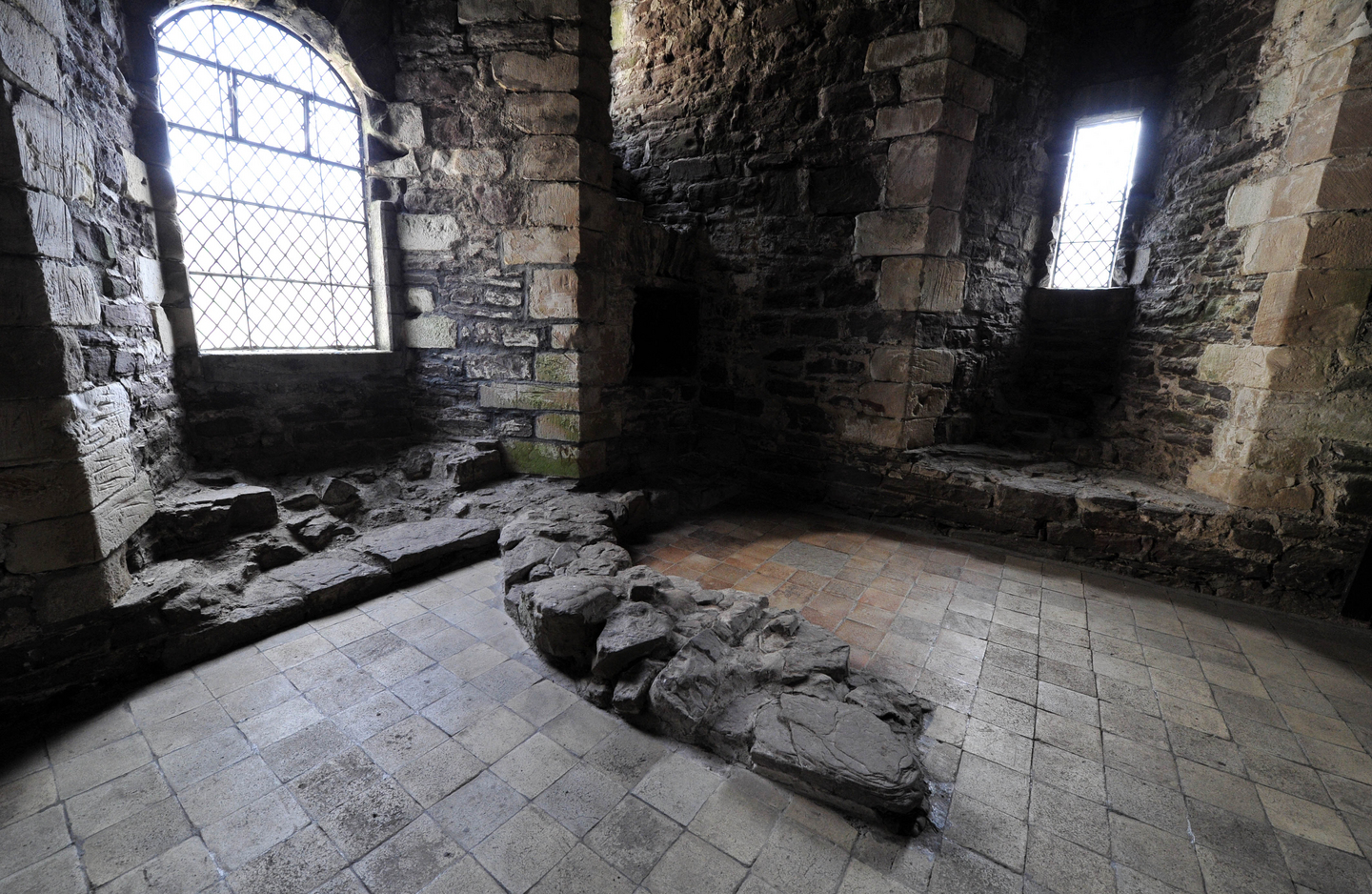 The interior to a historical building. The walls are stone, with arches framing two windows with light coming through them. There is the remnants of a stone wall on the floor. 