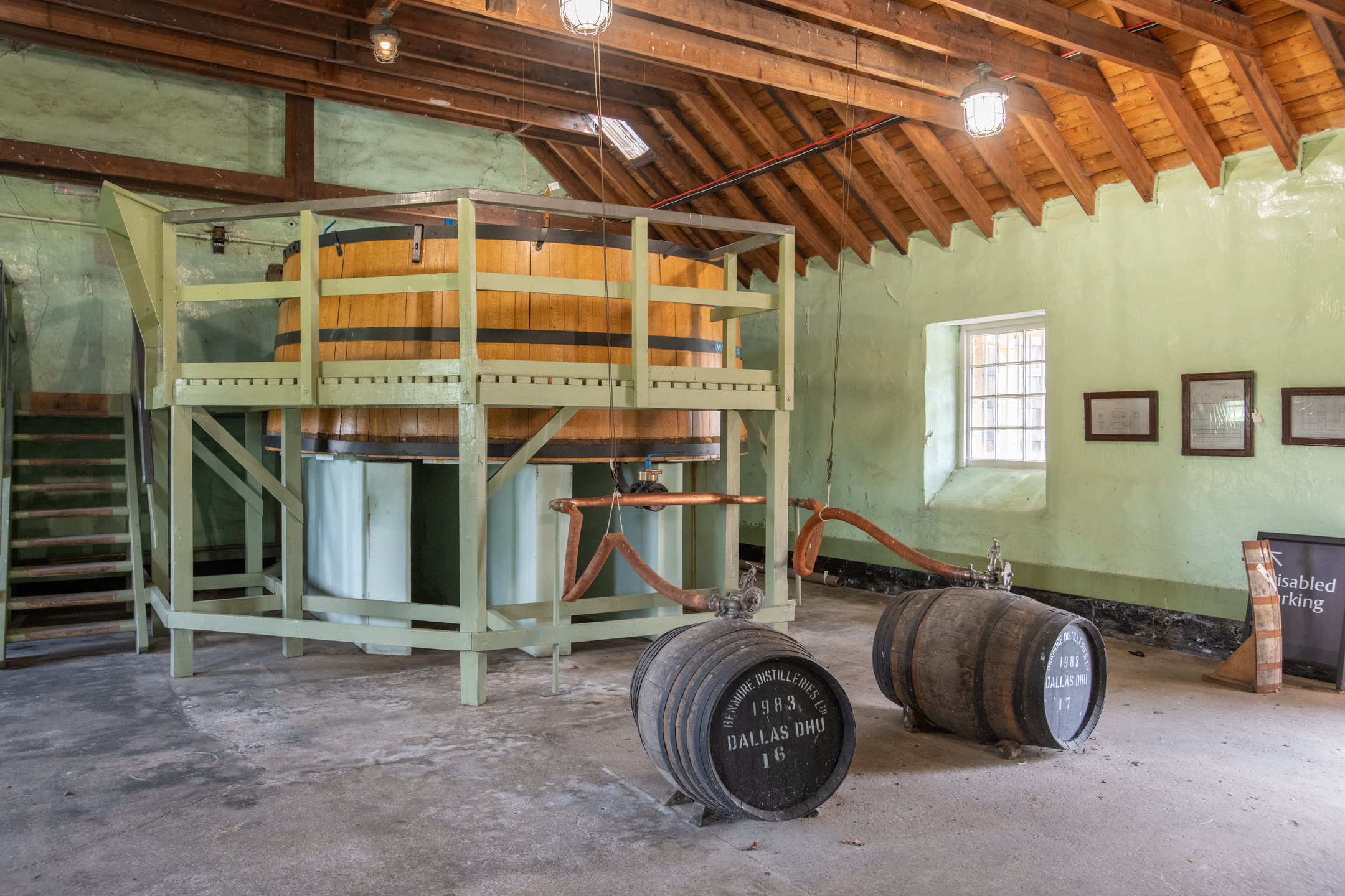 Historic wooden washback inside Dallas Dhu distillery, with copper pipework and whisky casks displayed beneath exposed timber roof beams.
