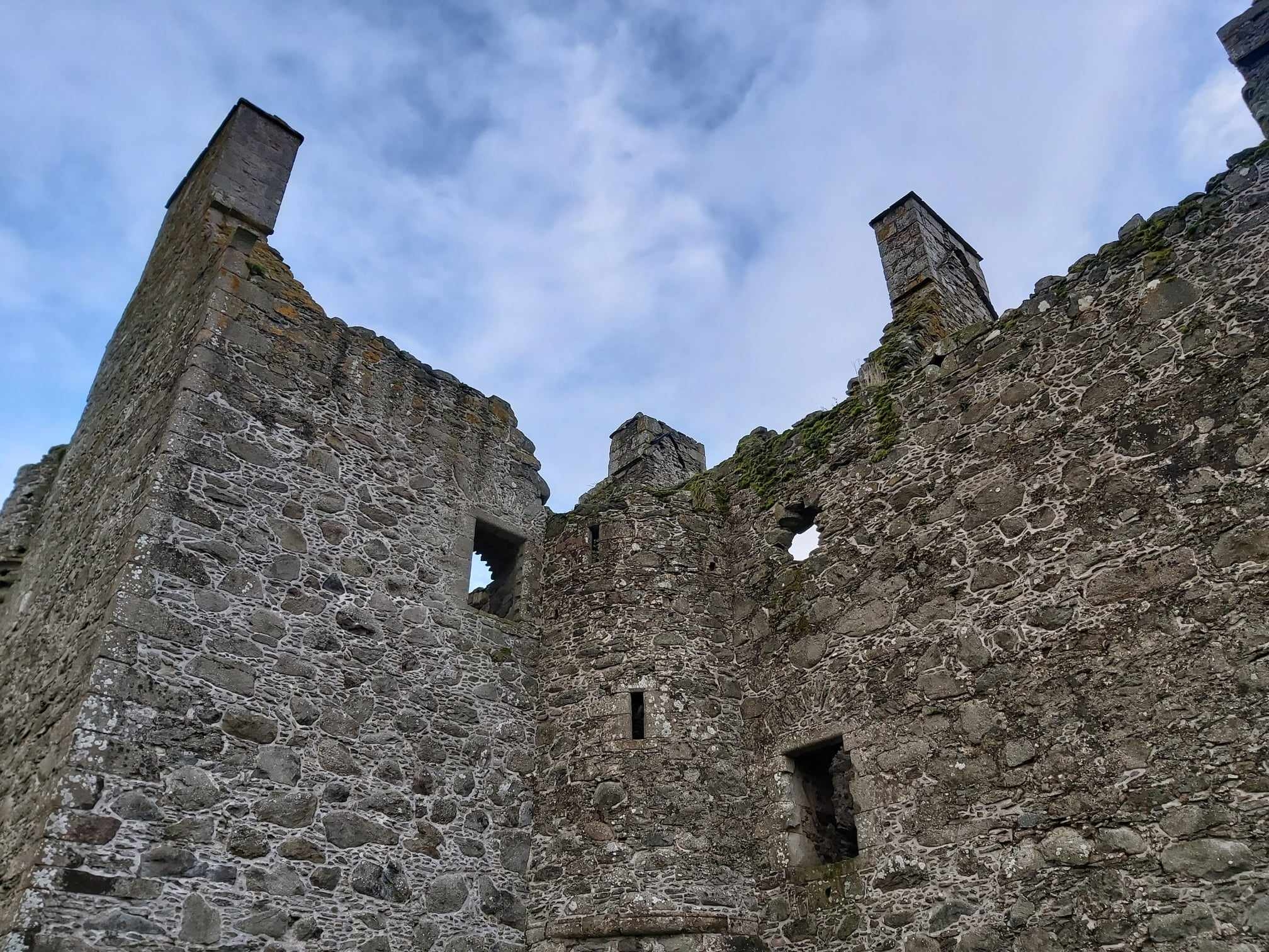 View of the ruined stone walls and tall chimneys of Glenbuchat Castle against a blue sky, showing weathered masonry and empty window openings.