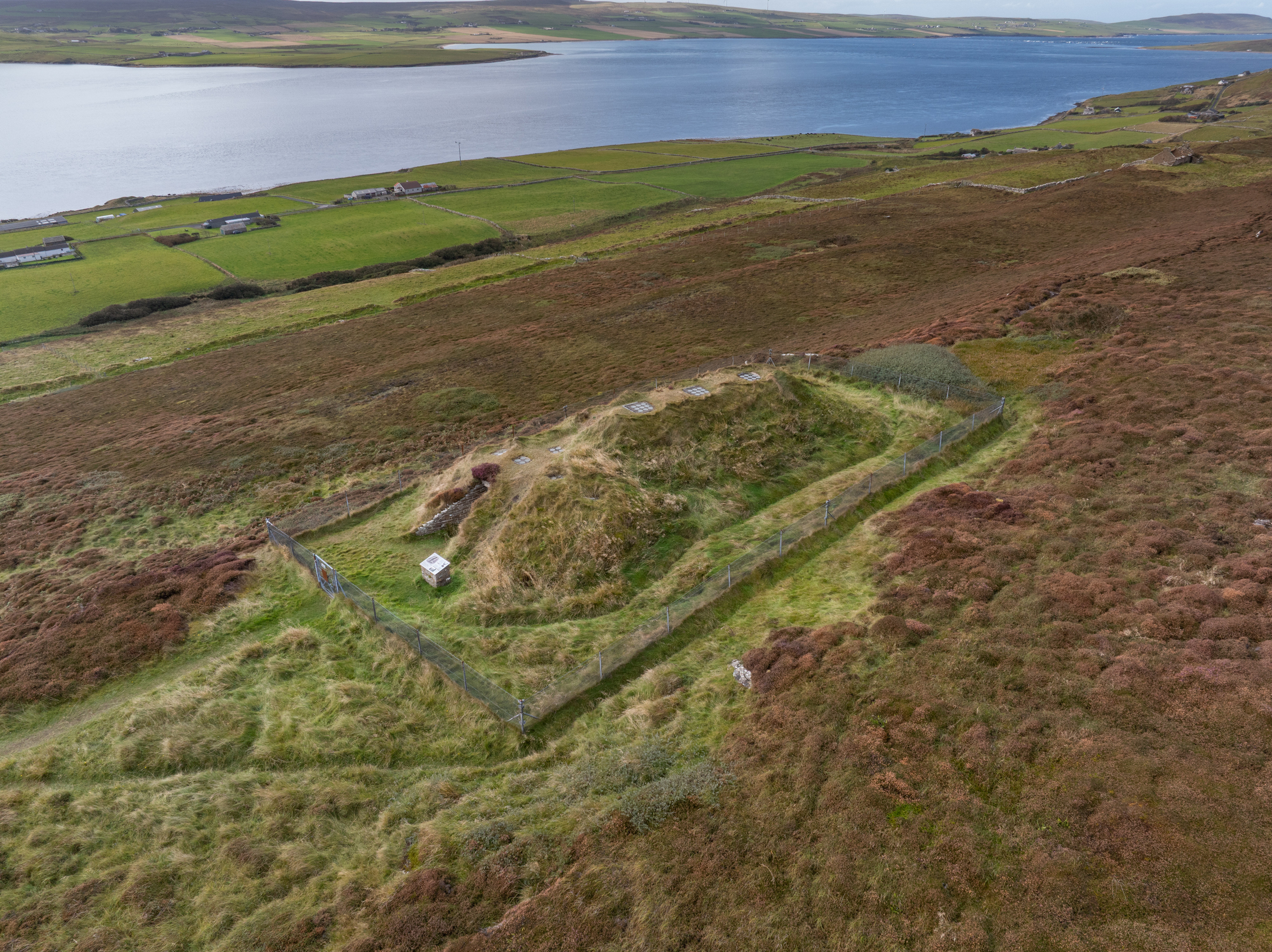 An aerial view of a chambered cairn. It is a vaguely oval-shaped grass covered mound surrounded by a fence. The surrounding island landscape features heather-covered moorland, agricultural land and a calm sea or sea inlet. 