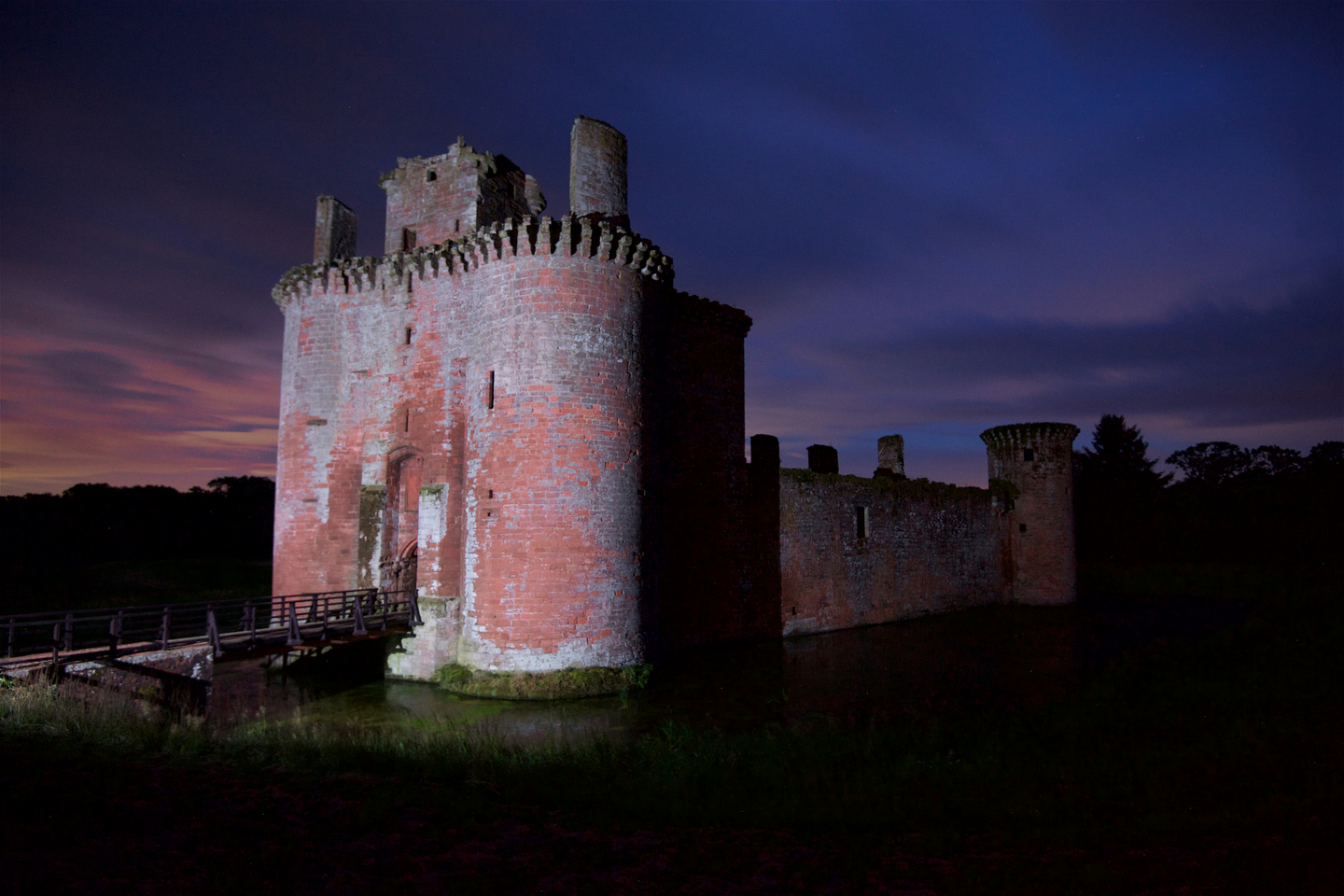 A red‑brick castle with round towers is lit against a dark night sky, with a wooden bridge crossing the surrounding water.