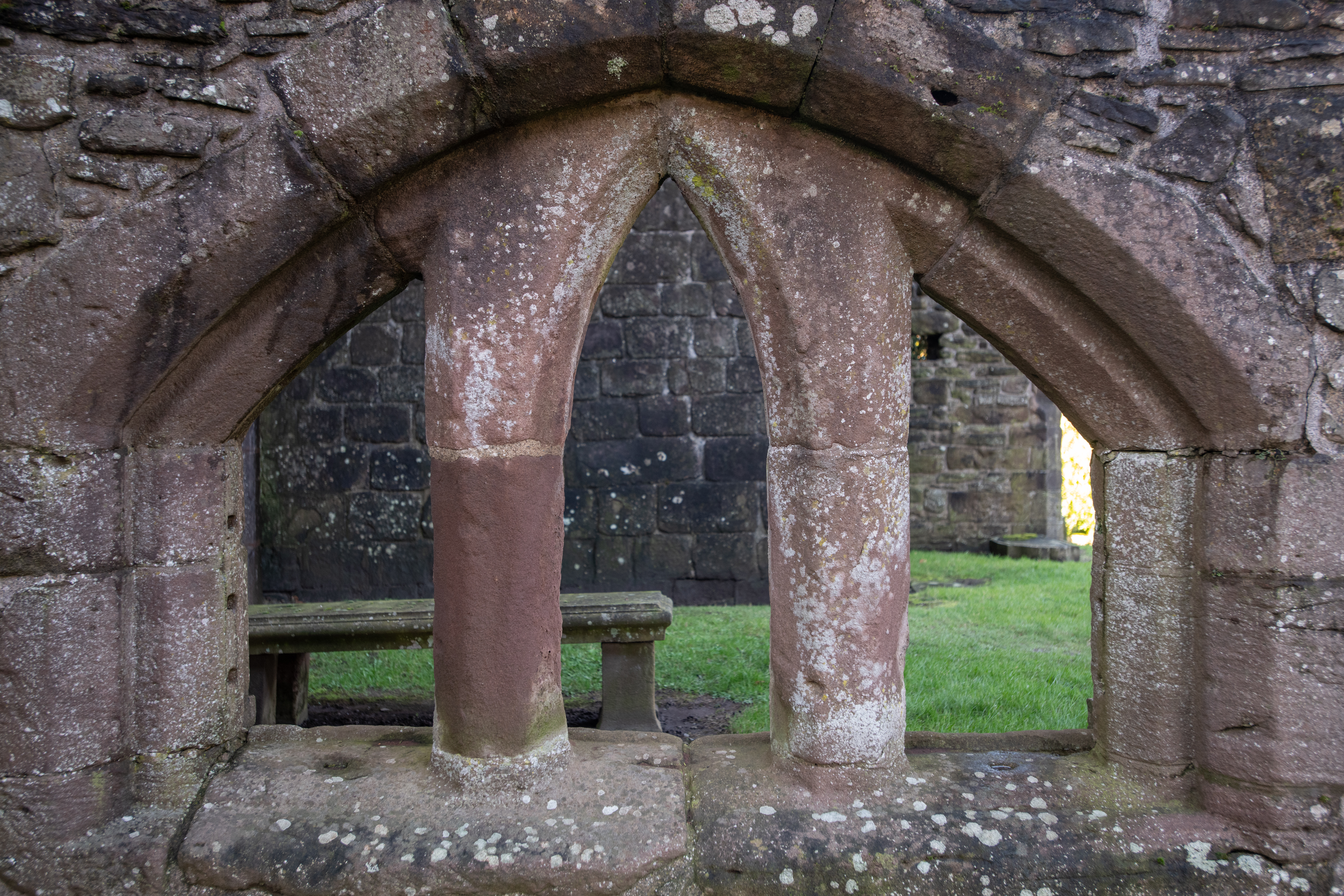A close-up of a double-arched stone window opening with weathered red sandstone columns, looking into the grassy interior of the ruins.