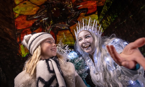 A child with blonde hair dressed in a winter hat, scarf and coat stands next to a woman dressed as an ice queen. The ice queen is holding out her hand to the camera and her and the child are looking at each other and smiling. She is dressed in a silver dress with a silver crown and silver hair. Her crown and dress is lit up with blue lights. In the background, there are red and yellow images projected onto a stone castle wall.