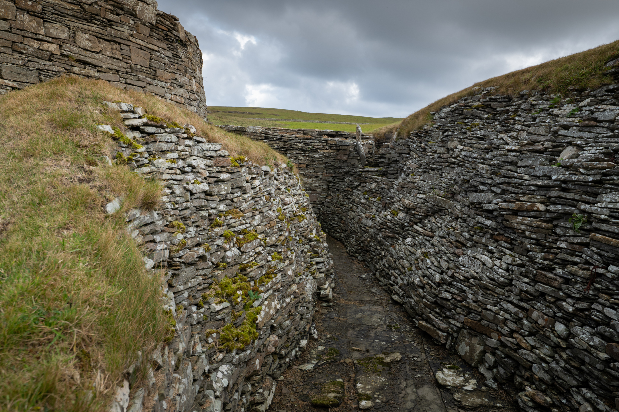 Narrow, stone-lined passageway beside the broch, with layered drystone walls rising on each side and grass covering the tops.