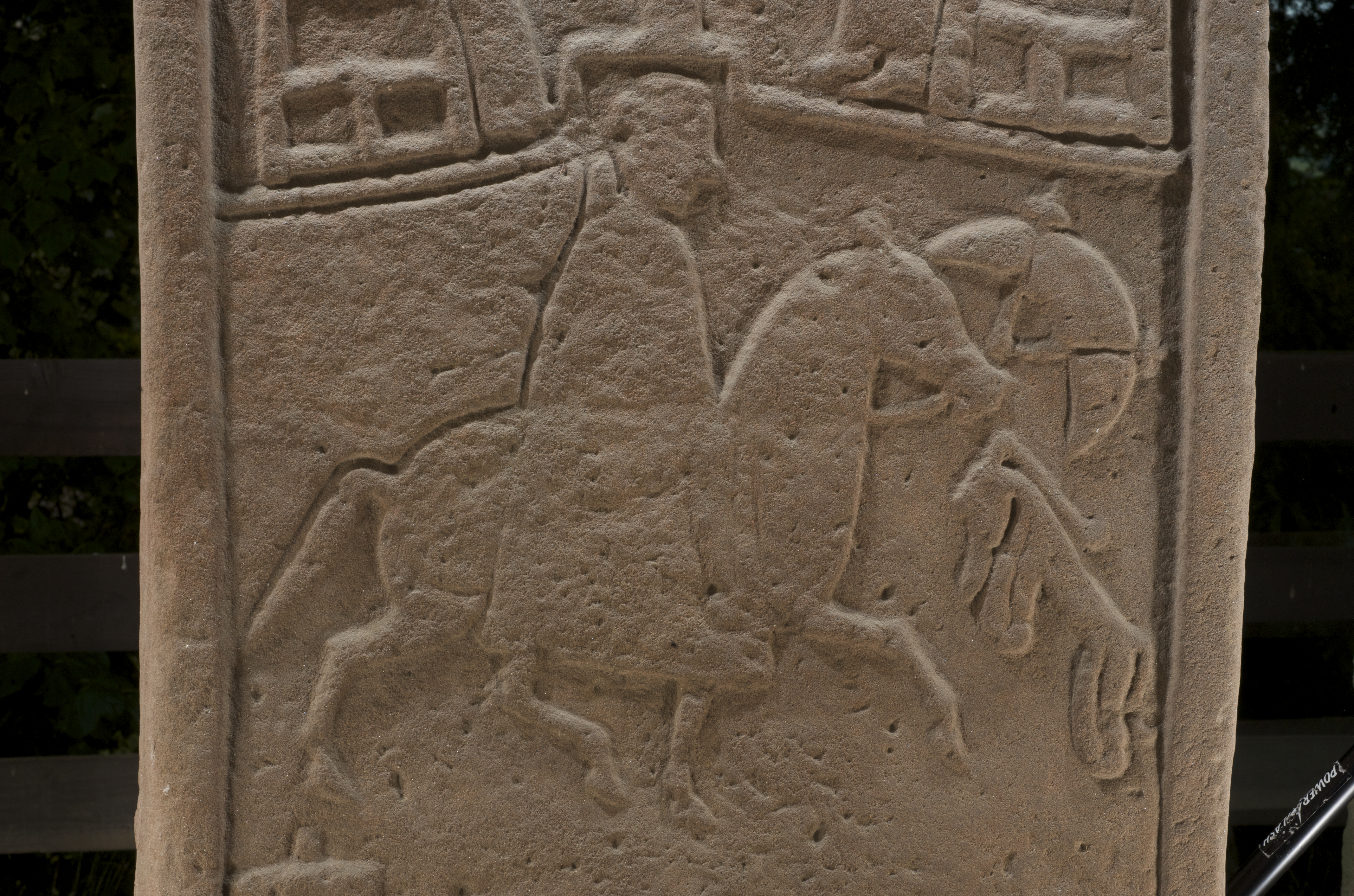 Detail of a mounted rider carved on the Dunfallandy Stone, shown in profile on horseback. The rider appears to hold a circular shield, and the horse is depicted with simple, linear features.