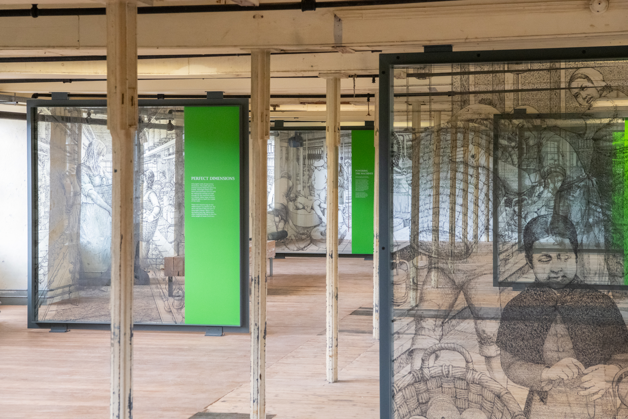A room filled with large framed illustrations printed on transparent screens, arranged in rows with green text panels between them in an open-plan gallery space.
