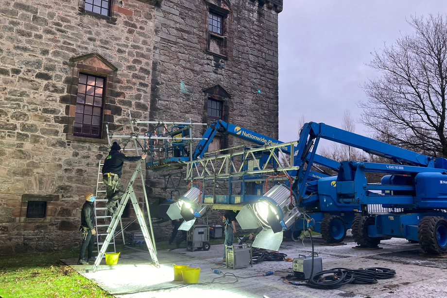 A large cherry picker in the grounds of a castle with large lighting, and people setting up to film at the castle