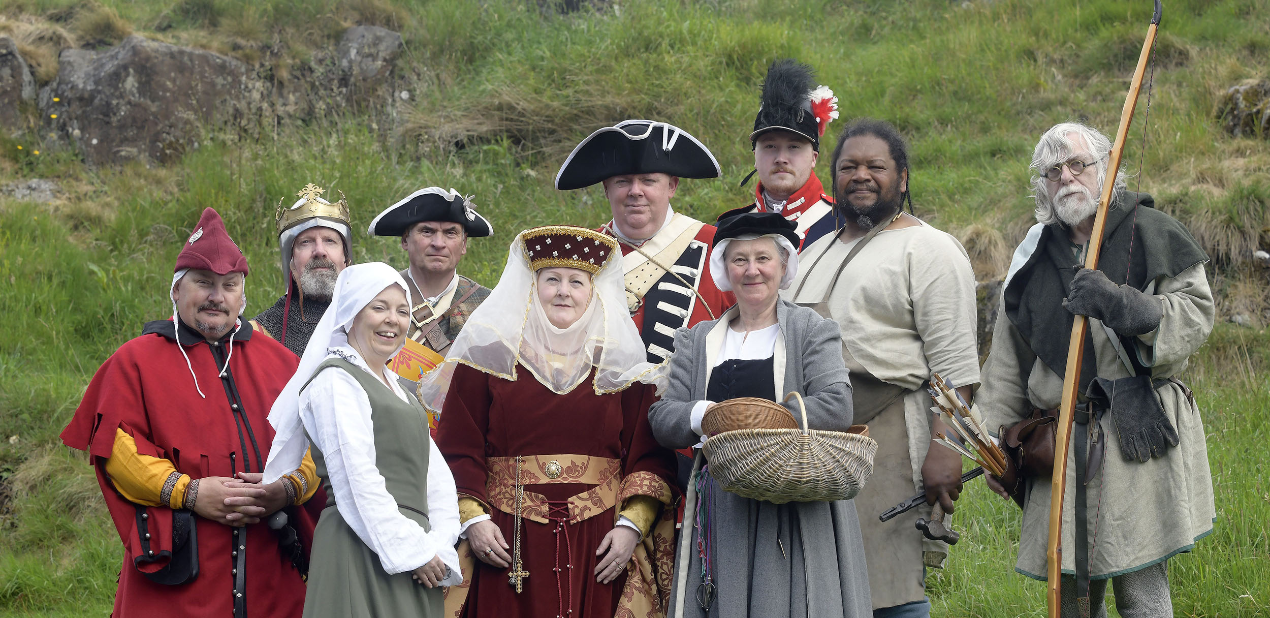 A group of costumed performers in different period costumes standing on a grassy hill 