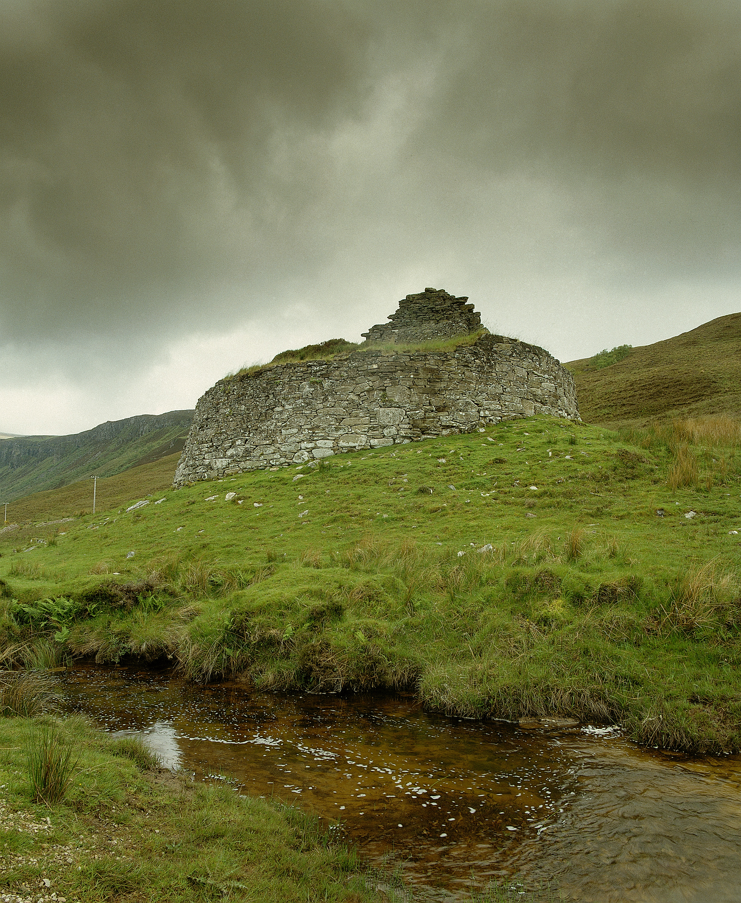 Rear view of the Dun Dornaigil broch, a circular drystone structure standing on a grassy slope beside a shallow, rocky stream. The broch’s upper walls are partially collapsed, forming an uneven silhouette against a sky filled with dark, heavy clouds. Rolling hills rise behind the ruin, creating a dramatic Highland landscape.