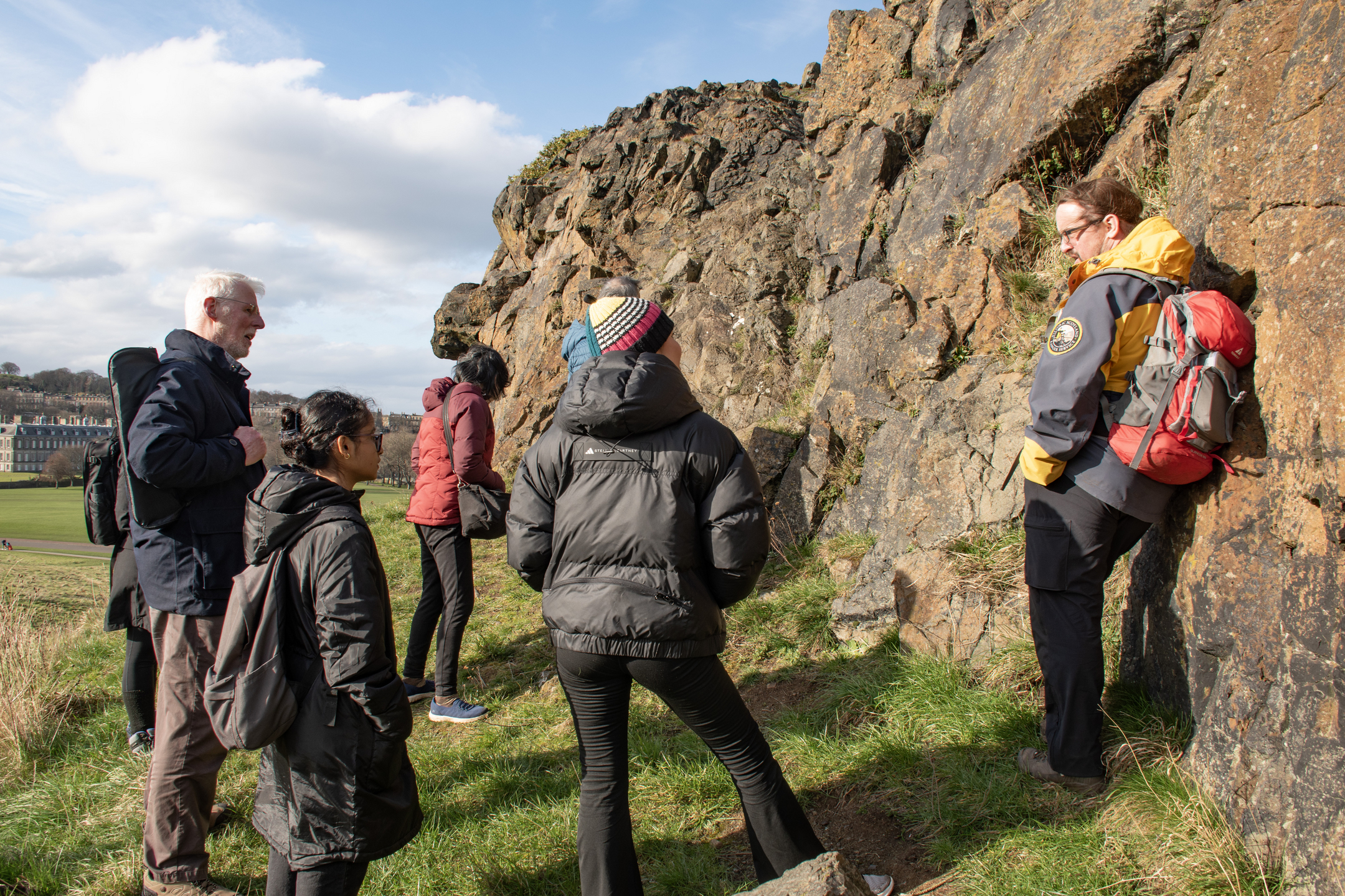 A HES ranger wearing an outdoor jacket and a rucksack stands beside a rocky crag and talks to a group of visitors about the geology in Holyrood Park.