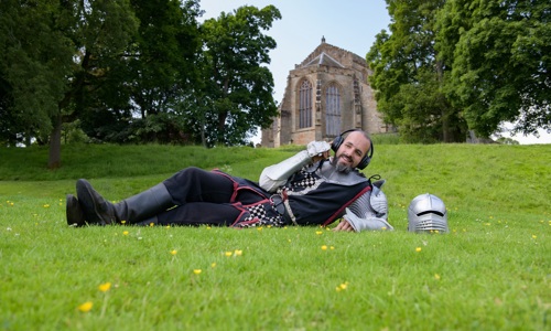 A man in medieval armour lies on the grass in front of the ruins of a castle in a comical pose with headphones on.