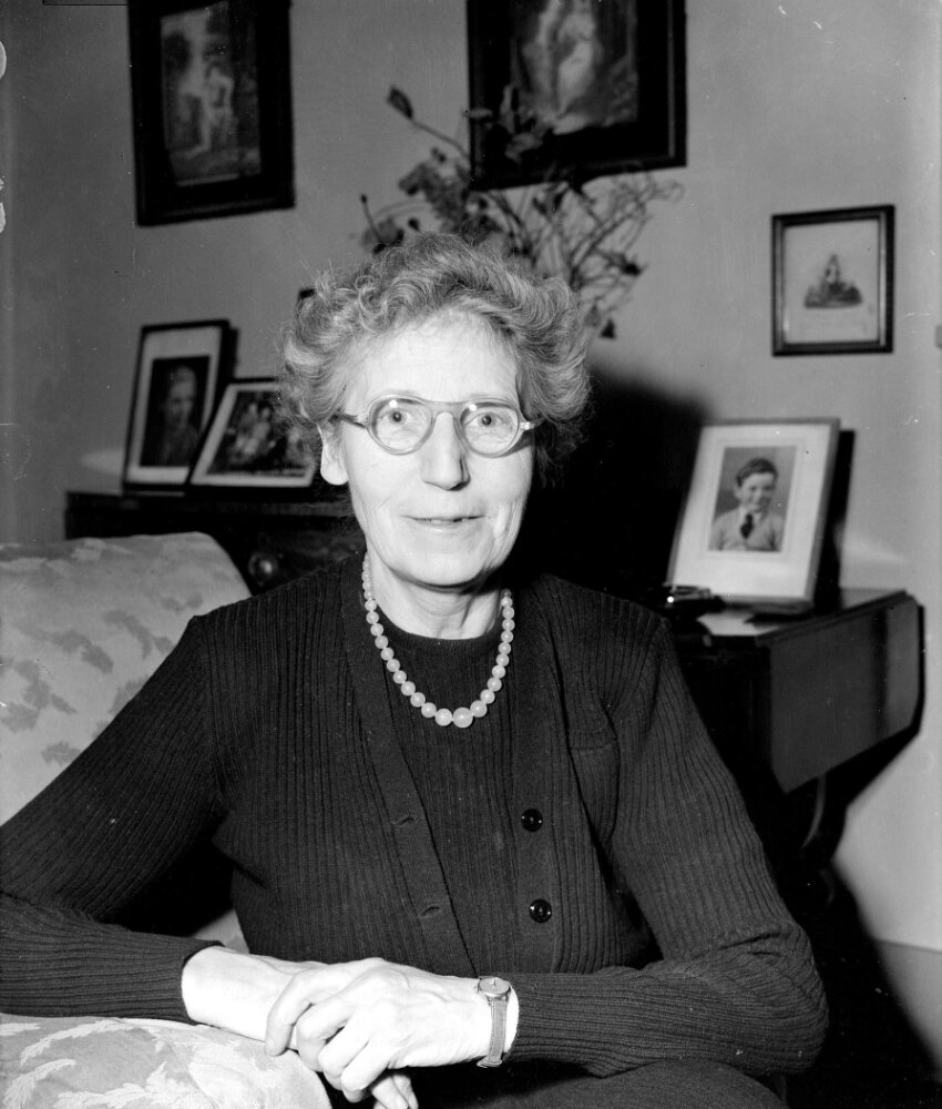 Black and white portrait photograph of a person sitting with their arms folded in an armchair. They are wearing glasses and a necklace, and behind them on a sideboard table are family photographs and a bunch of flowers in a vase.