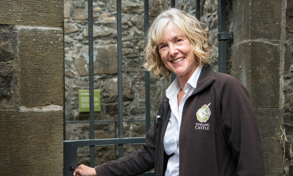 A lady with blonde hair, wearing a white shirt and an open black jacket with a Stirling Castle badge on the right-hand side is looking and smiling at the camera while holding a key inside the lock of a metal gate door inside a bricked castle building.