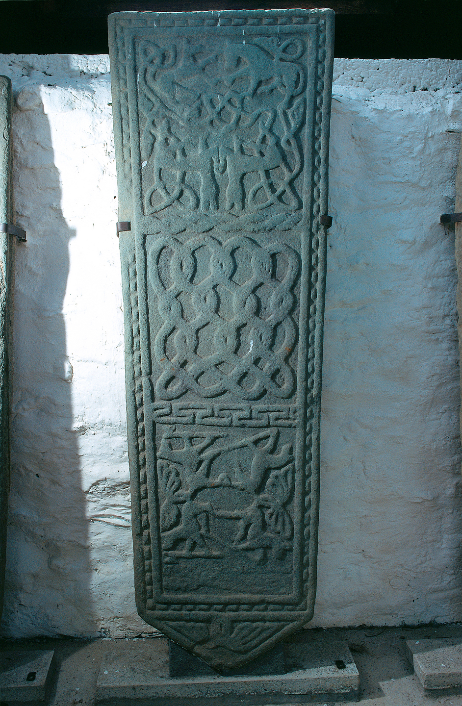 A sculptured stone leaning against a wall. There are three parts to the sculpting.