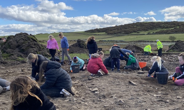 Children in jackets are knelt and digging in earth, with large buckets to the side of them. Some adults stand over them.