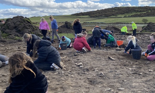 Children in jackets are knelt and digging in earth, with large buckets to the side of them. Some adults stand over them.