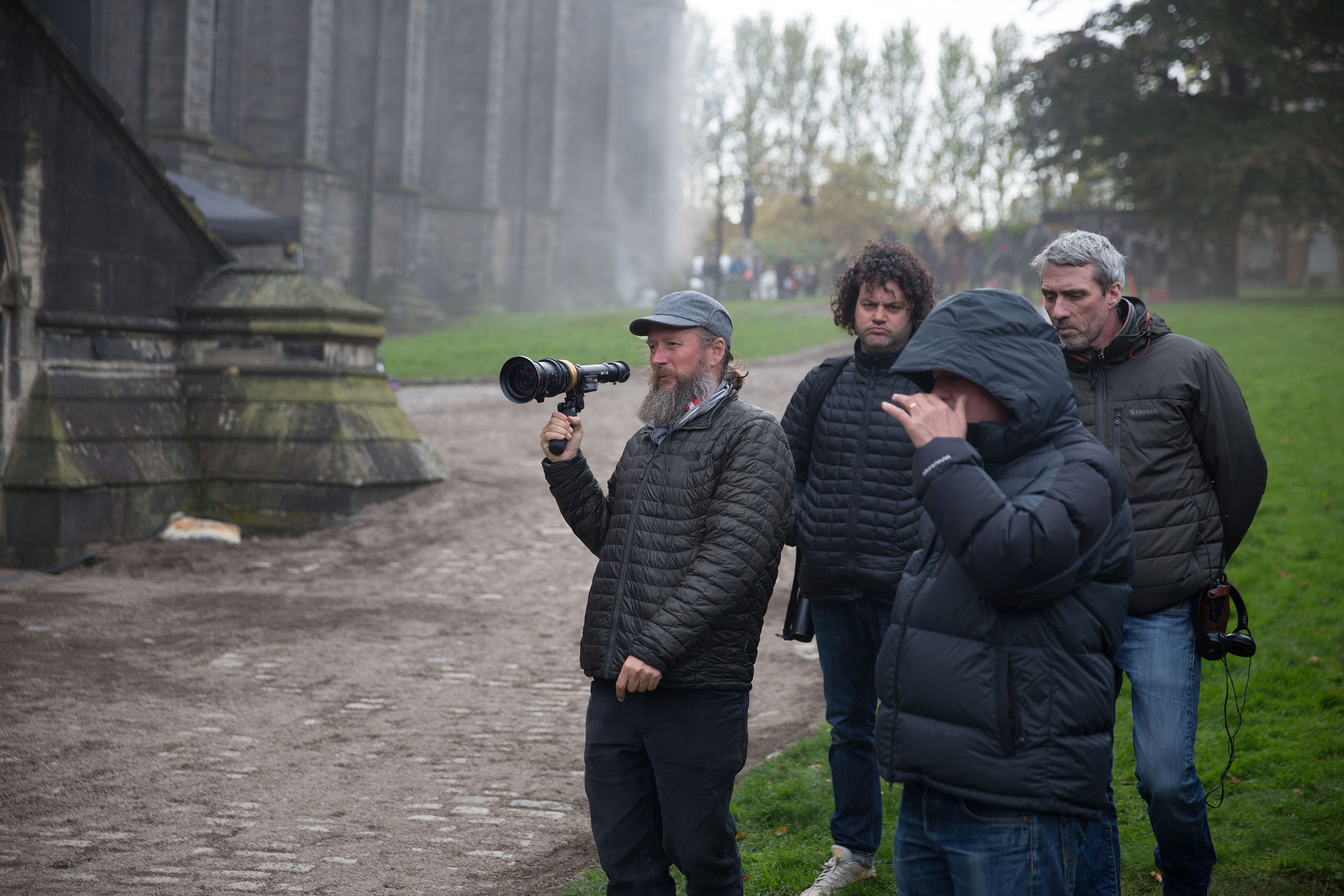 A film crew stands outdoors near a large stone building, with a cameraman holding a handheld rig as others watch the scene unfold.