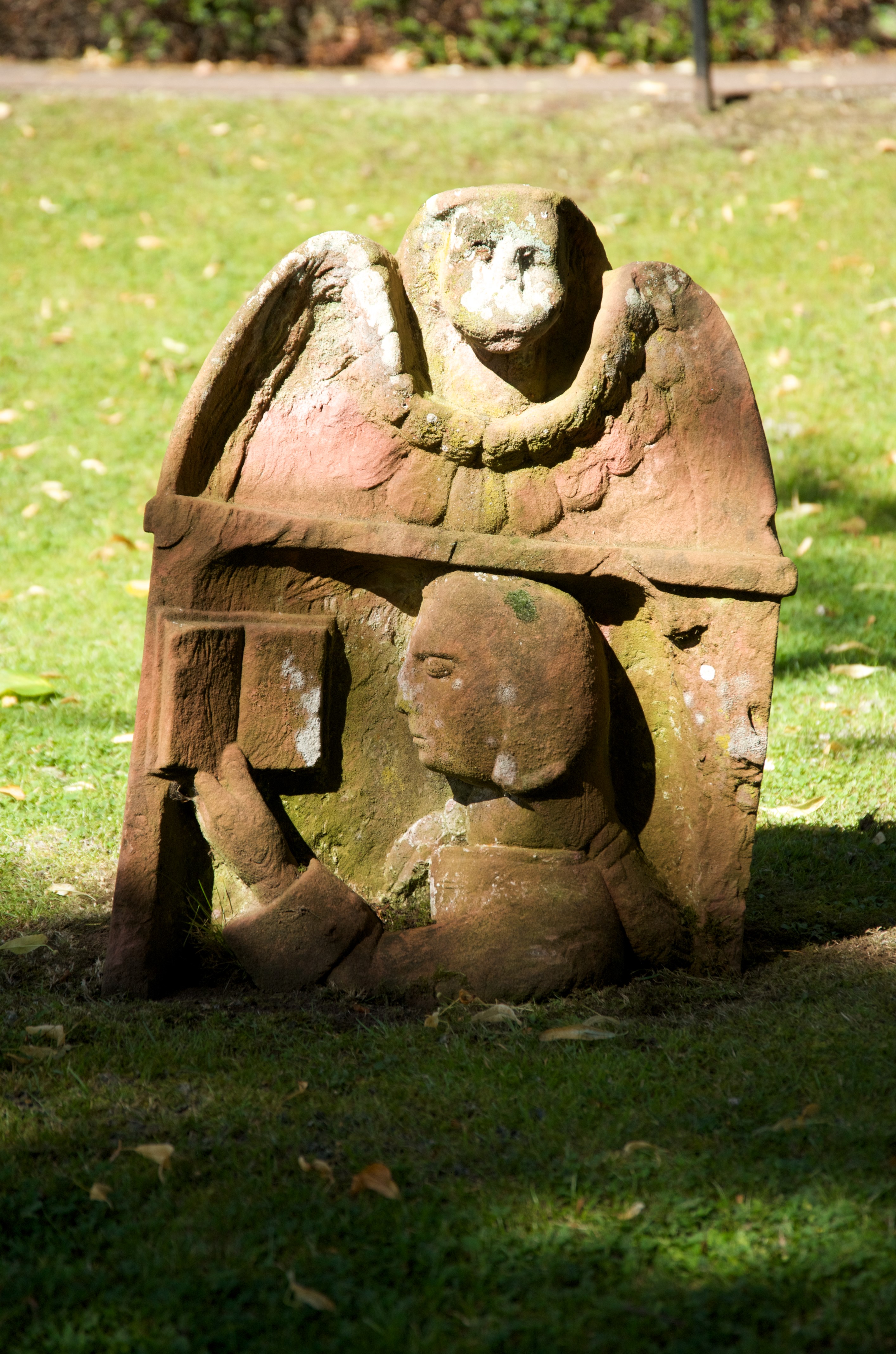 A small gravestone, with an unusual and striking stone carving of a person holding an open book, with an animal above their head. It is a short gravestone, amongst grass. Sun shines on the gravestone. 