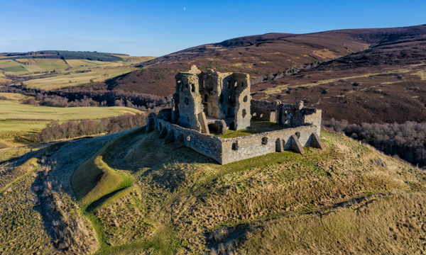 A ruinous stone castle stands atop a grassy hill. The castle is made up of a square roofless building with one wall missing and a number of holes where windows were. There is a wall forming a square around the outside of the building. There are rolling hills in the background and blue sky above.