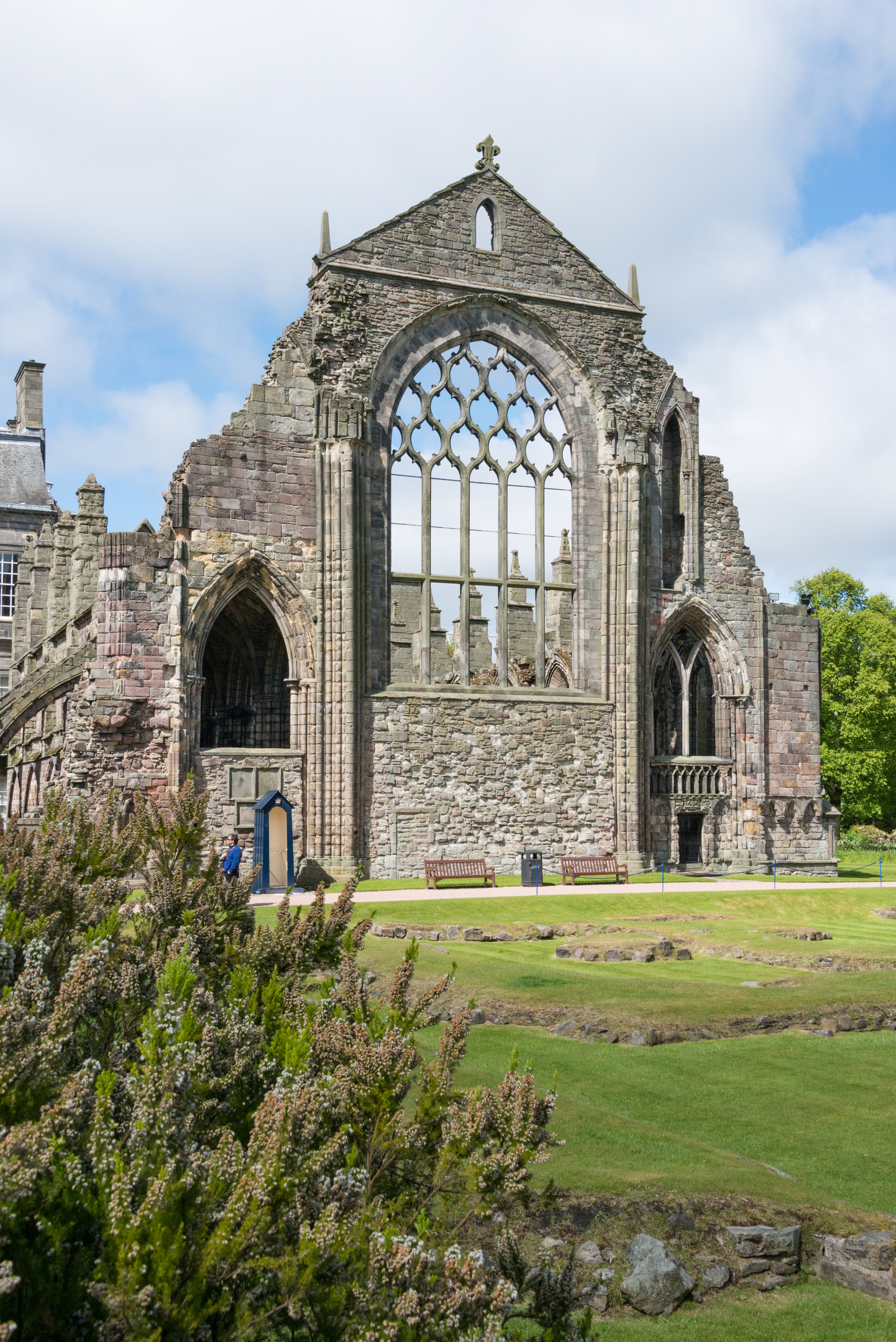 A view of the back wall of the nave, taken from outside the abbey.  The arched main window frame takes up most space on the wall but there are also smaller arched windows to either side. There are gardens which include old stone ruins in the ground in the foreground of the image.