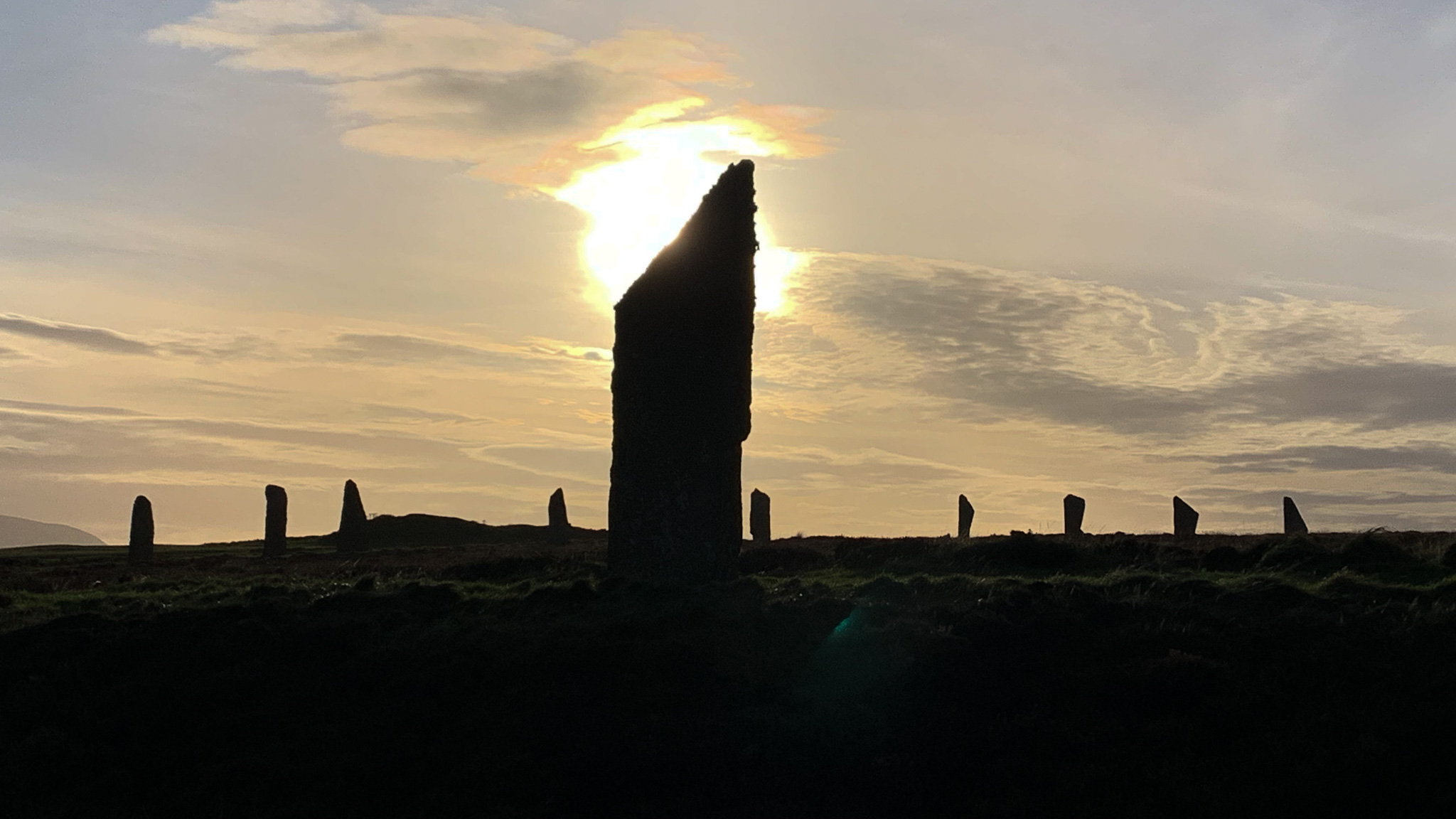 An atmospheric shot of a large standing stone in front of the sun. There is a line of other standing stones in the background, appearing smaller because they are further away.
