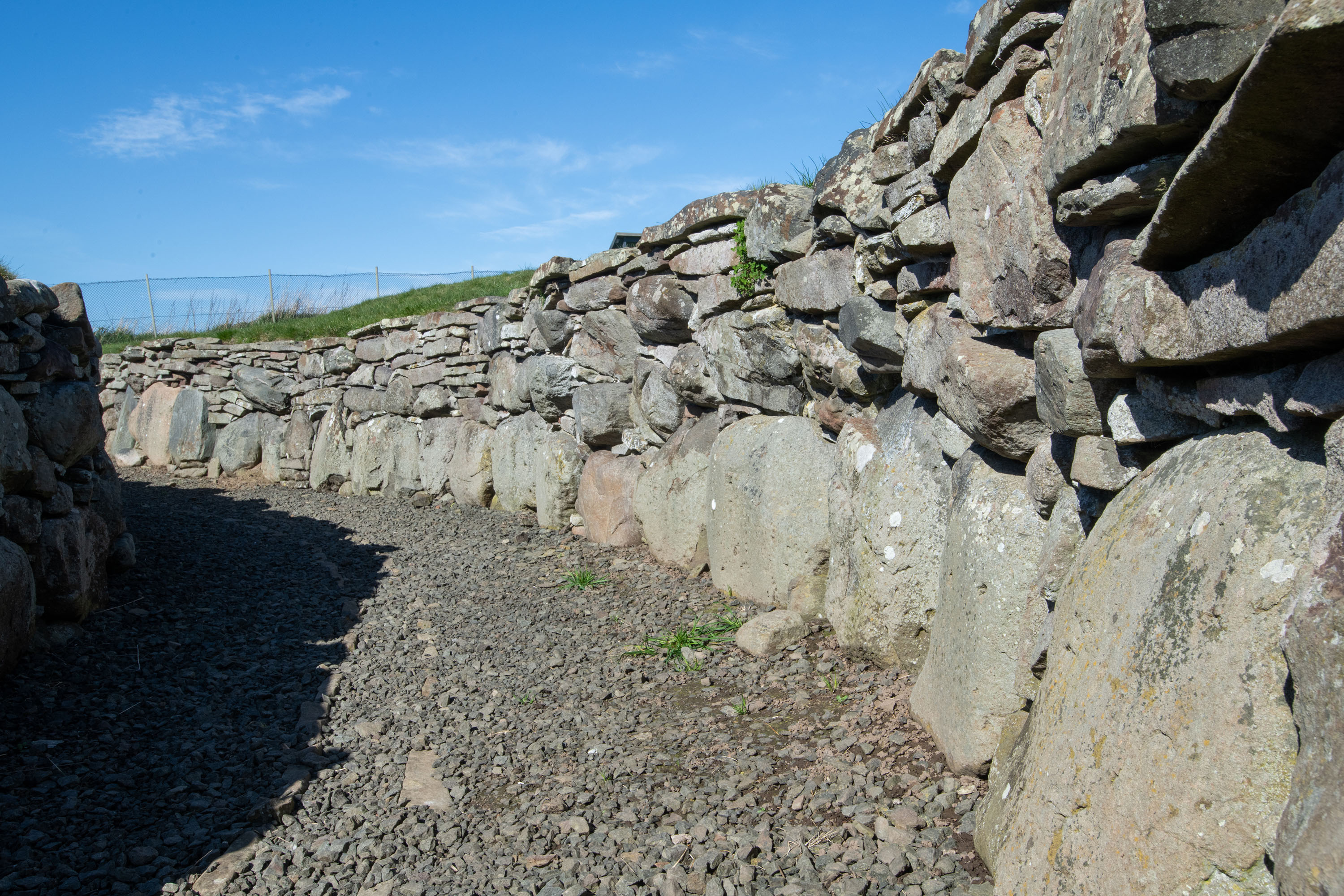 Close-up view inside Ardestie Earth House, showing a narrow passage lined with stacked dry‑stone walls. Large upright stones form the lower course, with smaller, irregular stones built above them. The floor is covered with loose gravel, and the walls curve gently under a bright blue sky.