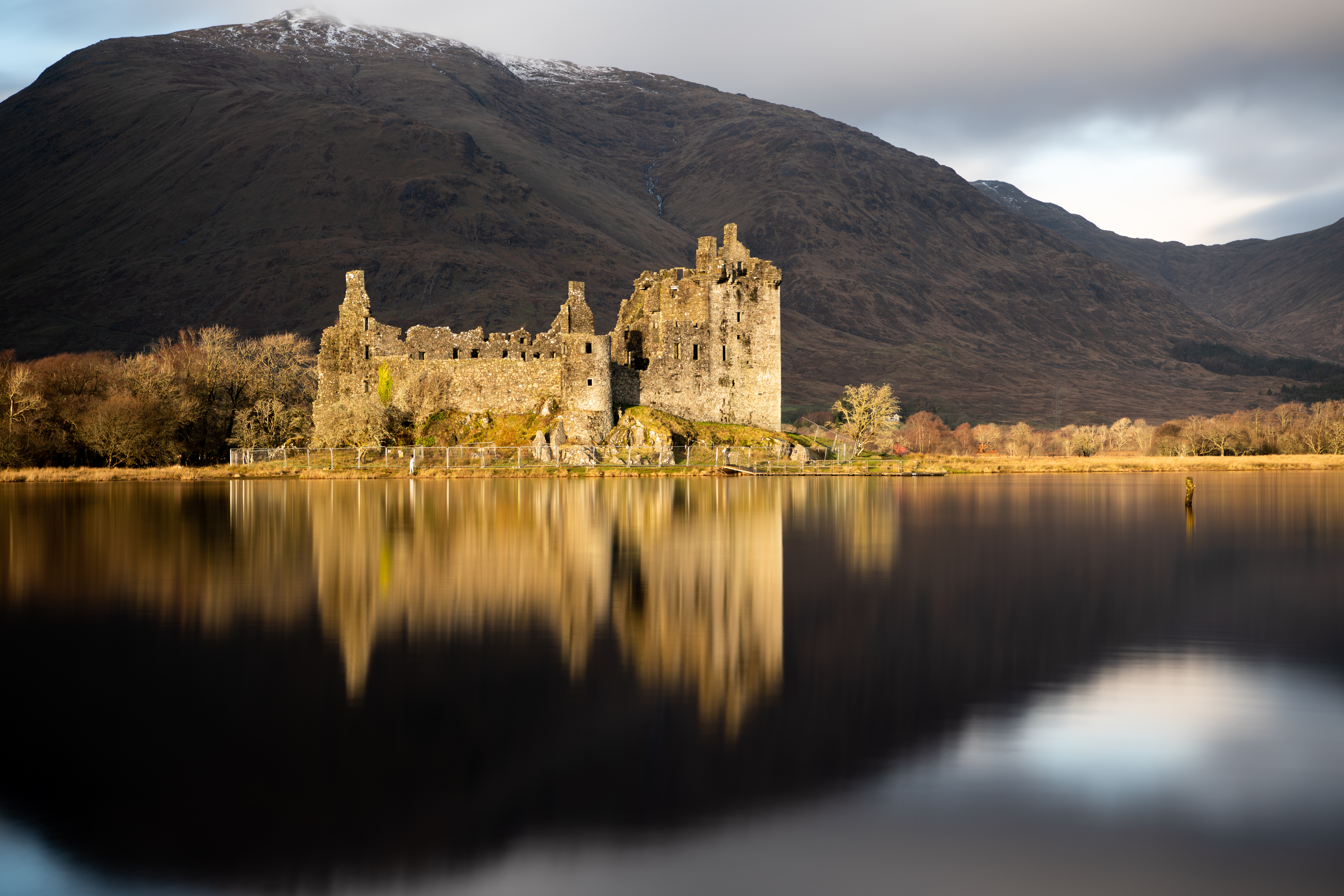 Sunlight shining on a castle in front of a loch, lighting it up in golden hues with a dark mountain looming large behind it