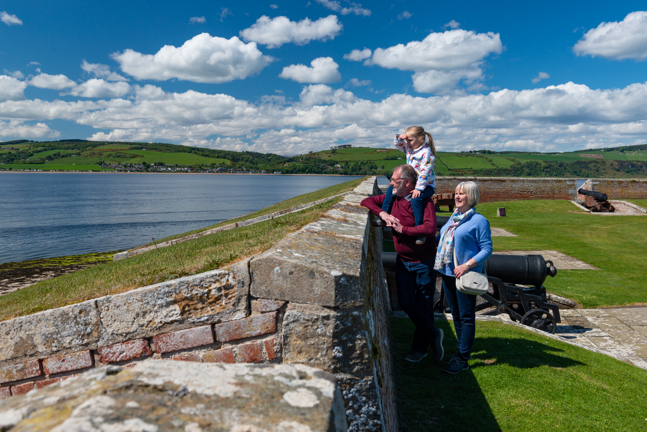 A family stands at the sea wall of Fort George, looking across the water with historic cannons positioned on the lawn behind them.