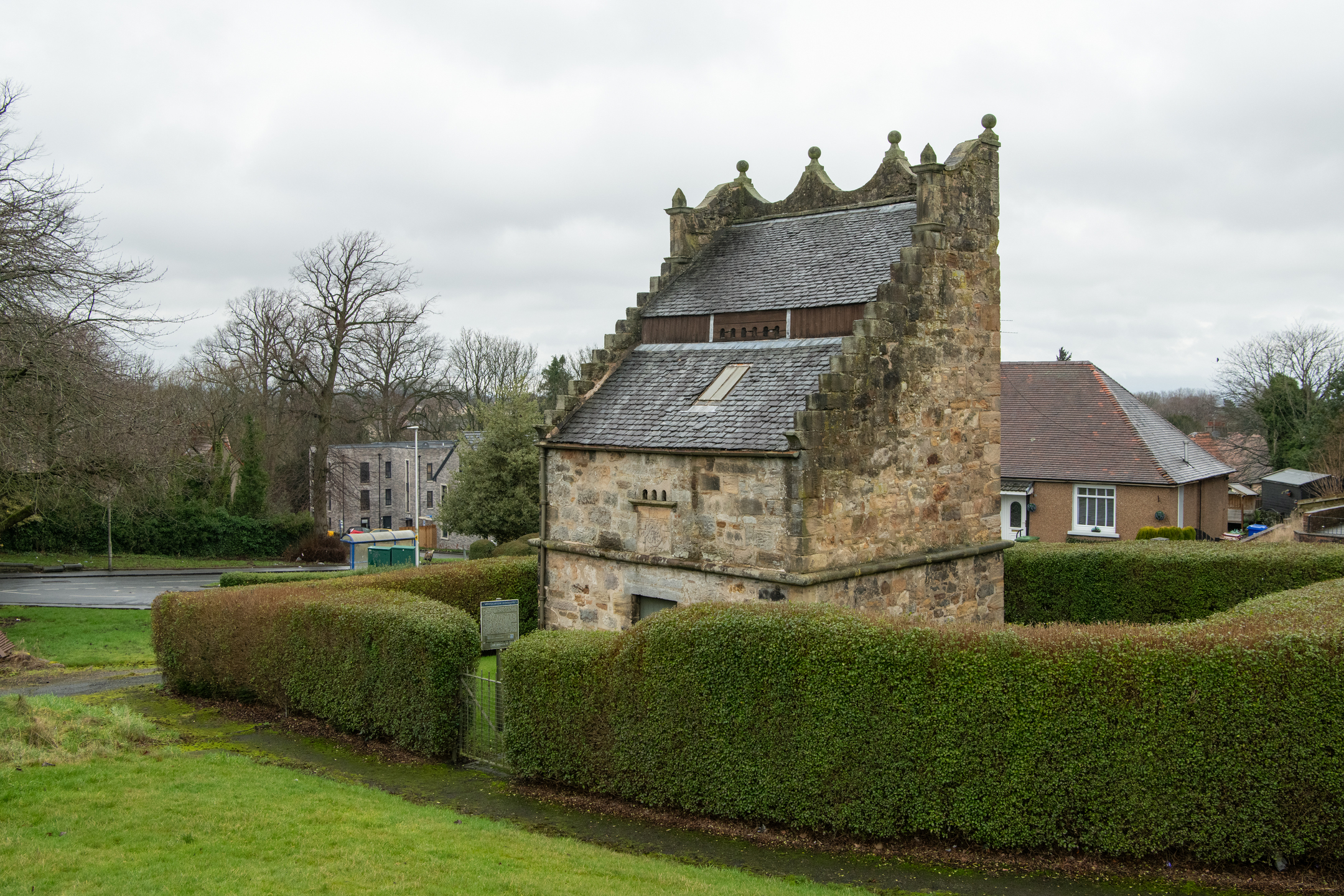 A historic stone dovecot with crow‑stepped gables and a slate roof, surrounded by neatly trimmed hedges, with houses and winter trees in the background under a grey sky.